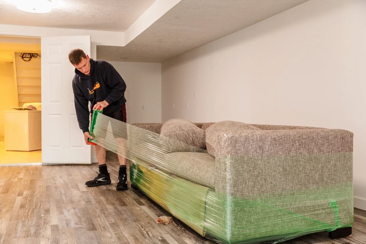 A man is wrapping a beige sofa with green plastic stretch wrap in a living room.