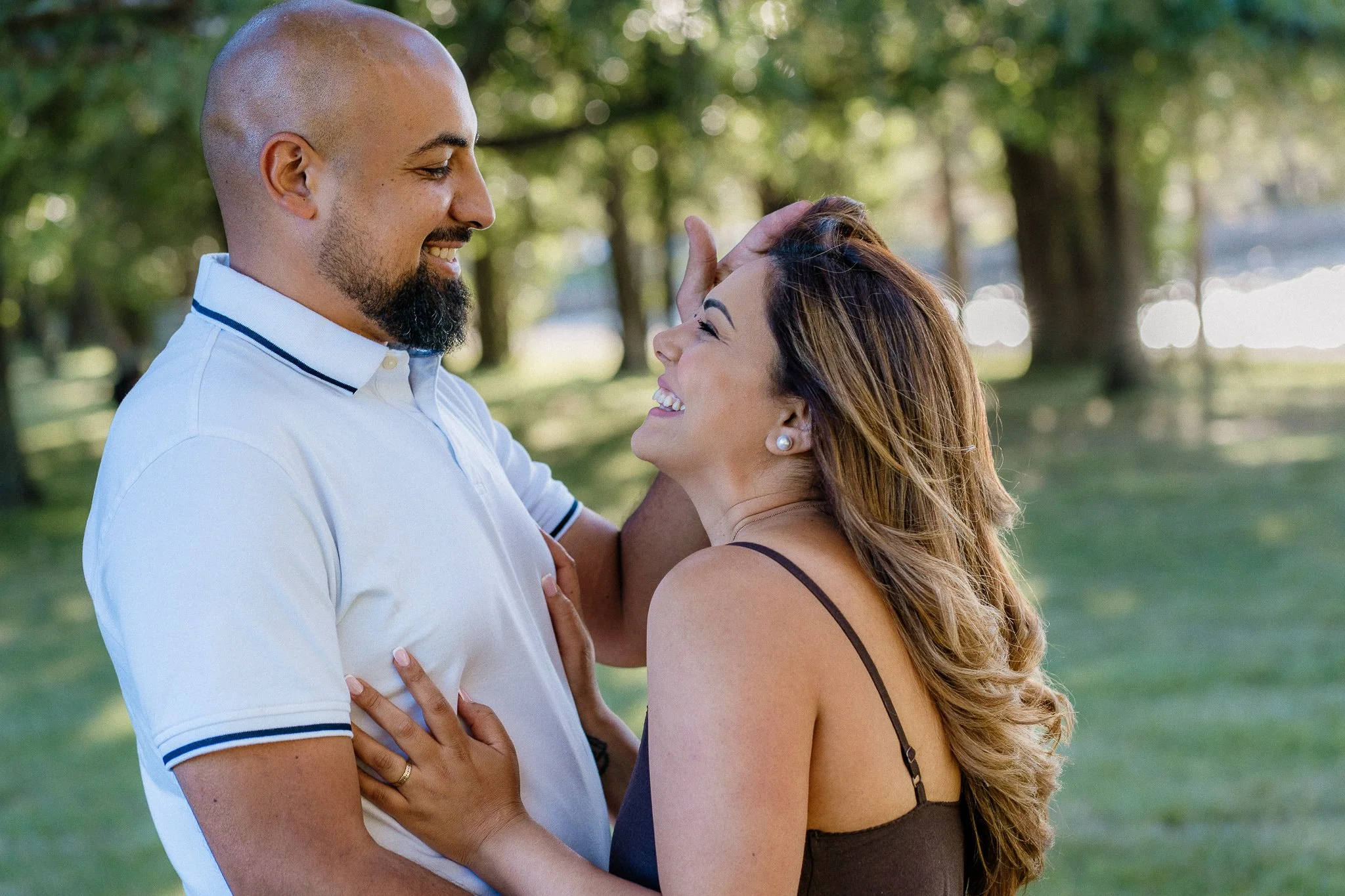 A man and a woman are smiling and sharing a moment of affection outdoors in a park with trees and sunlight in the background. The man has a beard, is bald, and is wearing a white polo shirt. The woman has long, wavy hair, is wearing earrings and a da