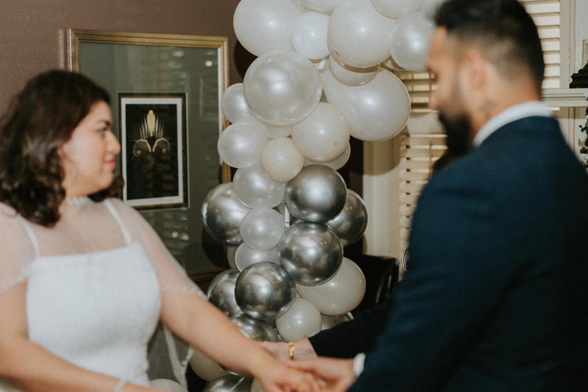 A woman and a man holding hands, standing in front of a cluster of white and silver balloons at what appears to be a celebration or event.