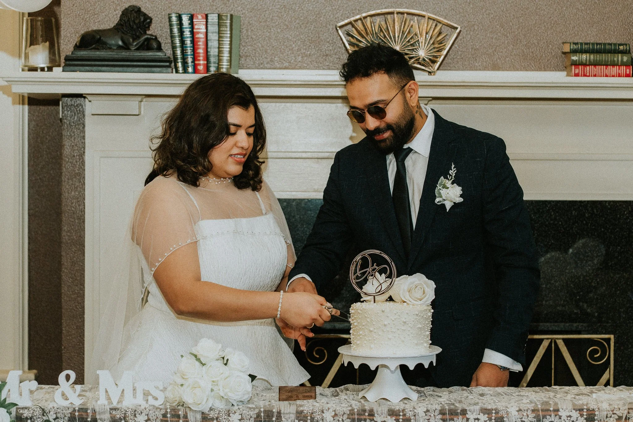 A bride and groom cutting a wedding cake together at their wedding reception, standing in front of a fireplace with books and decorative items on the mantel.