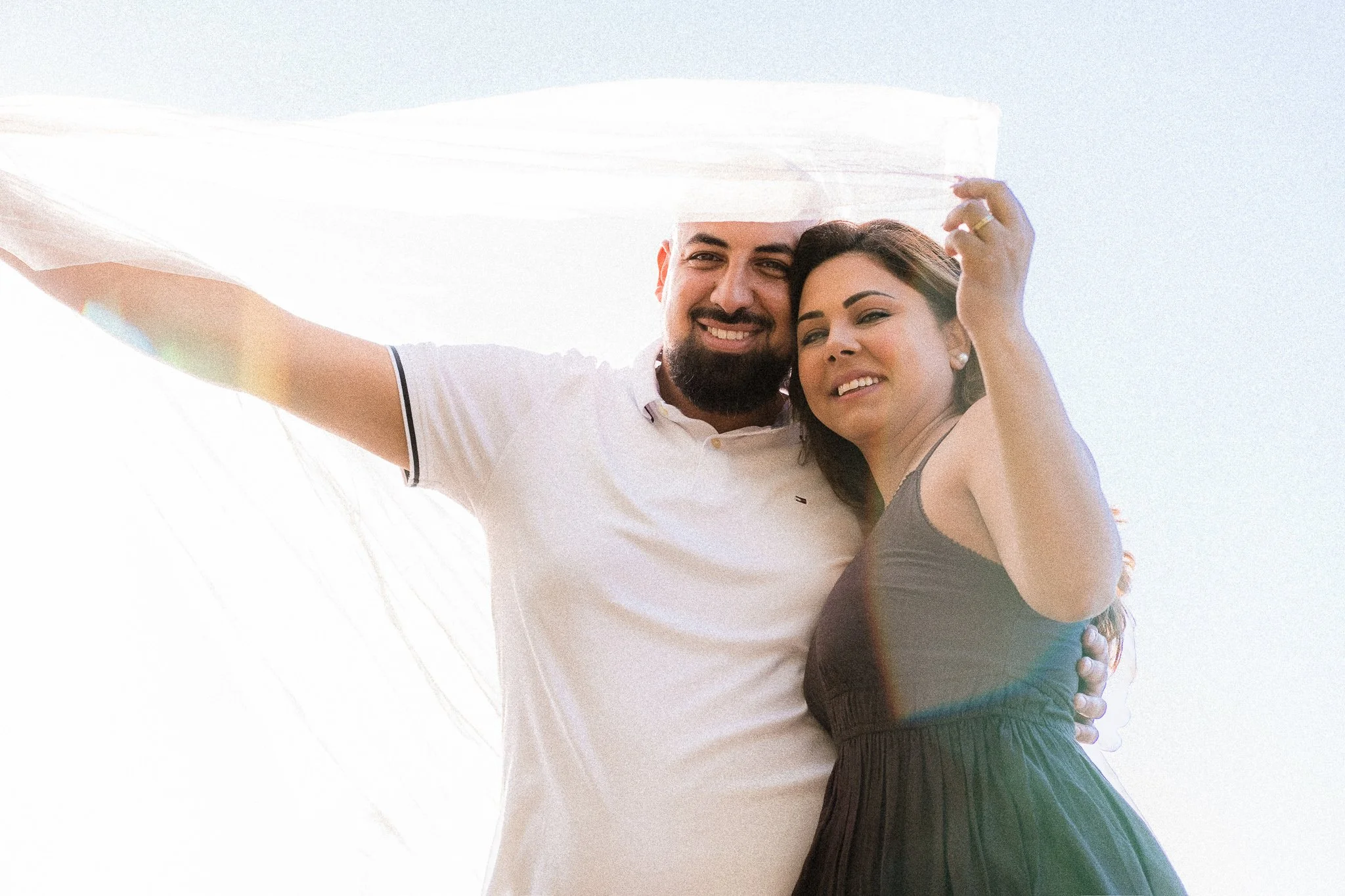 A smiling man and woman standing closely together, outdoors, with the woman holding a light-colored fabric or veil above their heads.