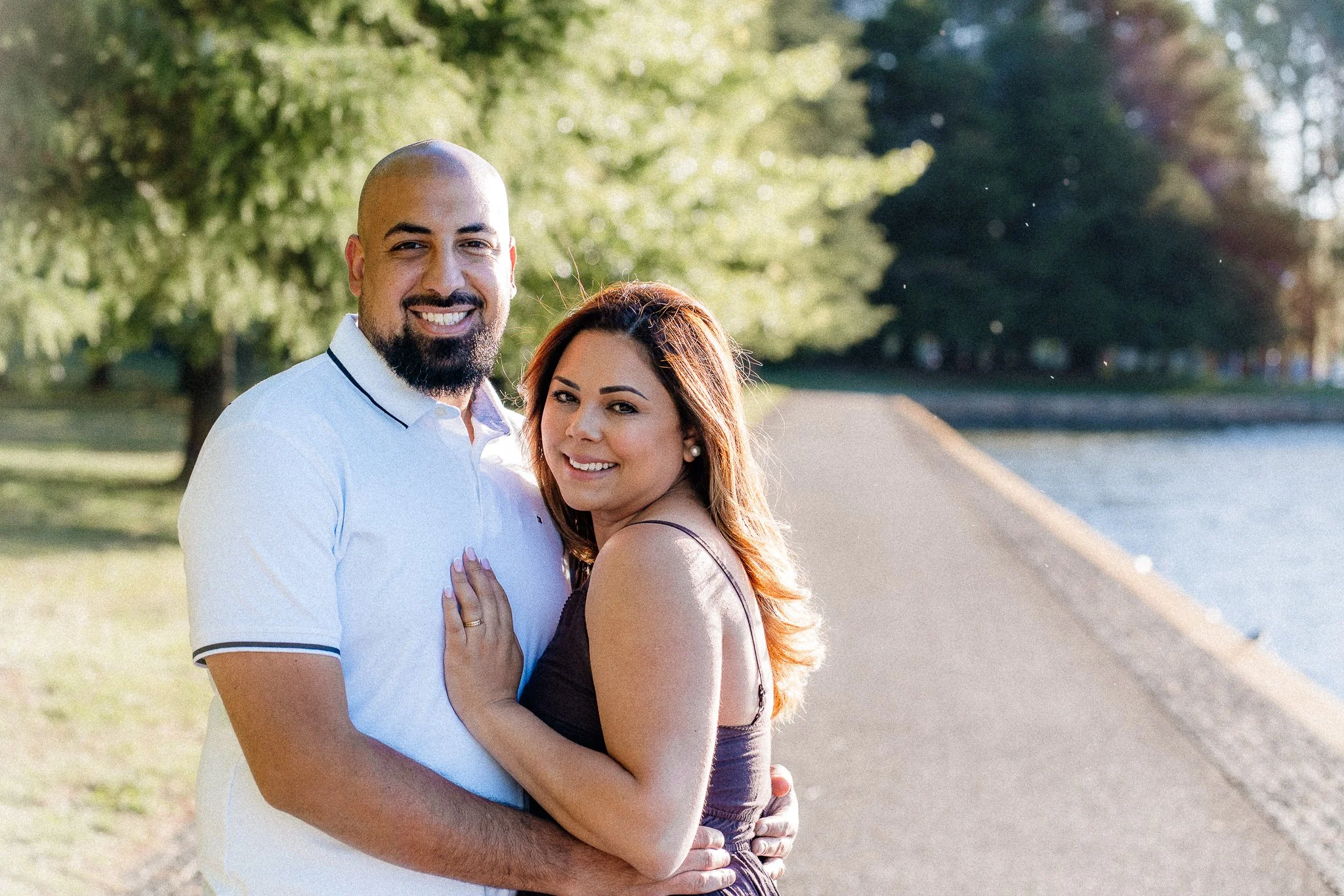 A smiling couple, a man with a bald head and beard dressed in a white polo shirt, and a woman with long hair in a black top, embracing outdoors on a pathway with trees and green foliage in the background.