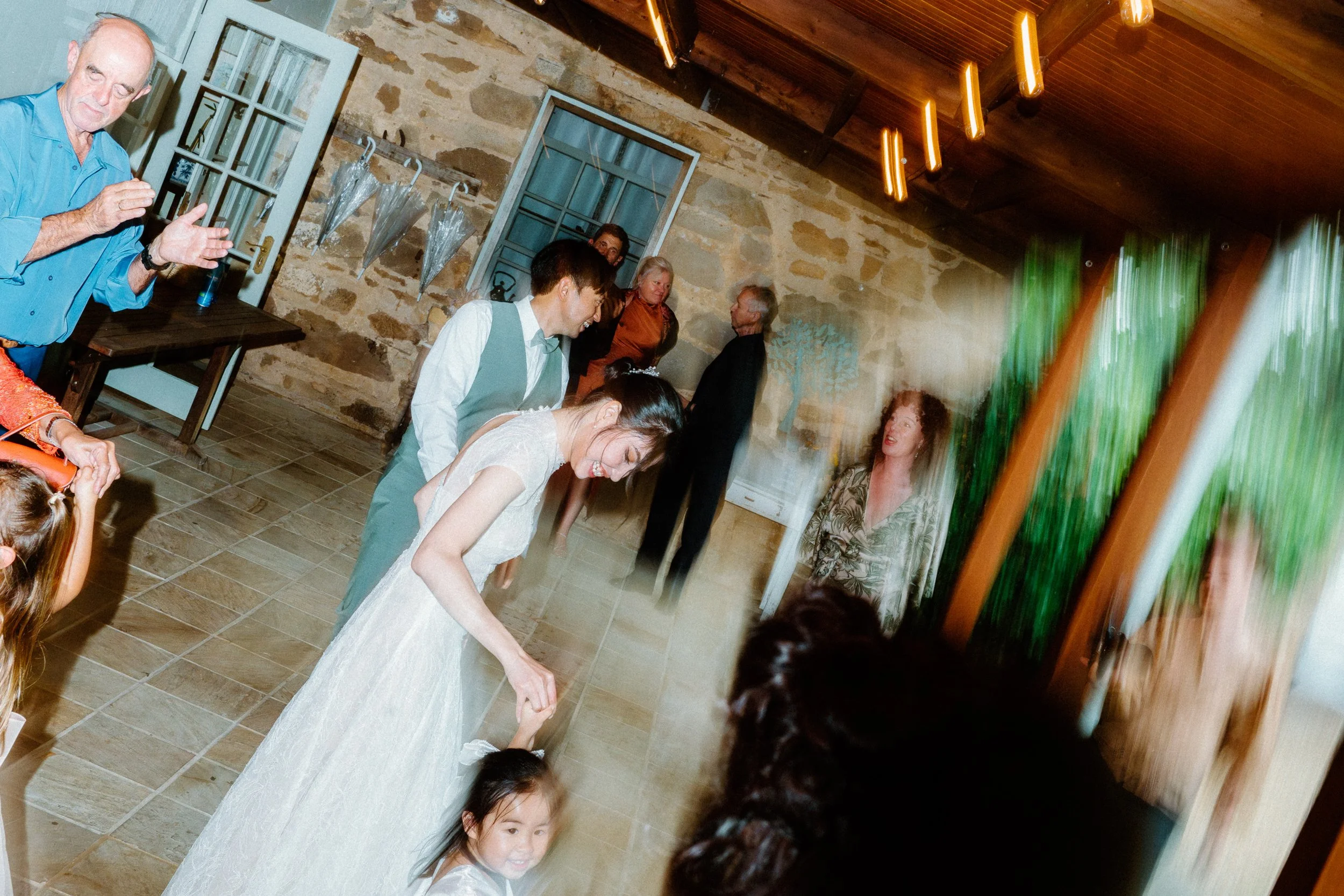 A group of people at a celebration, possibly a wedding reception, with a woman in a white dress and a young girl in a white dress in the foreground. Several adults are standing and smiling in the background, with a rustic stone wall and windows behin