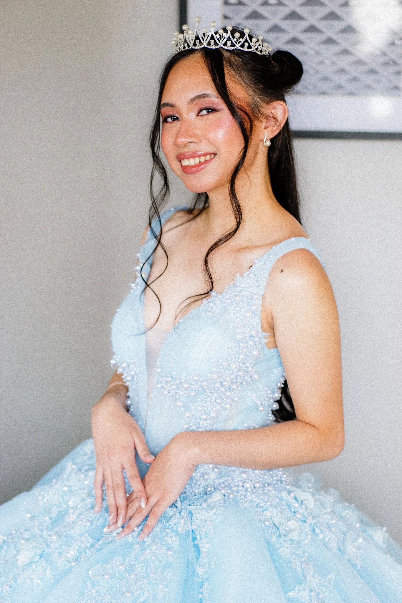 A young woman wears a tiara and a light blue dress decorated with pearls and beads, smiling and posing with her hands folded in front of her.