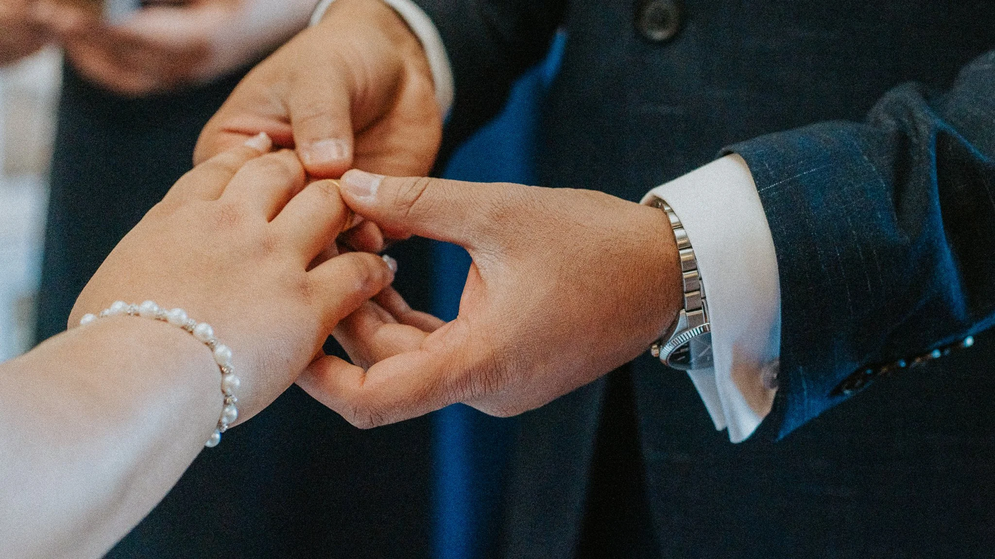 Close-up of a man and woman holding hands, with the woman's hand decorated with a pearl bracelet. The man is wearing a suit with a white shirt, a watch, and a dark tie.