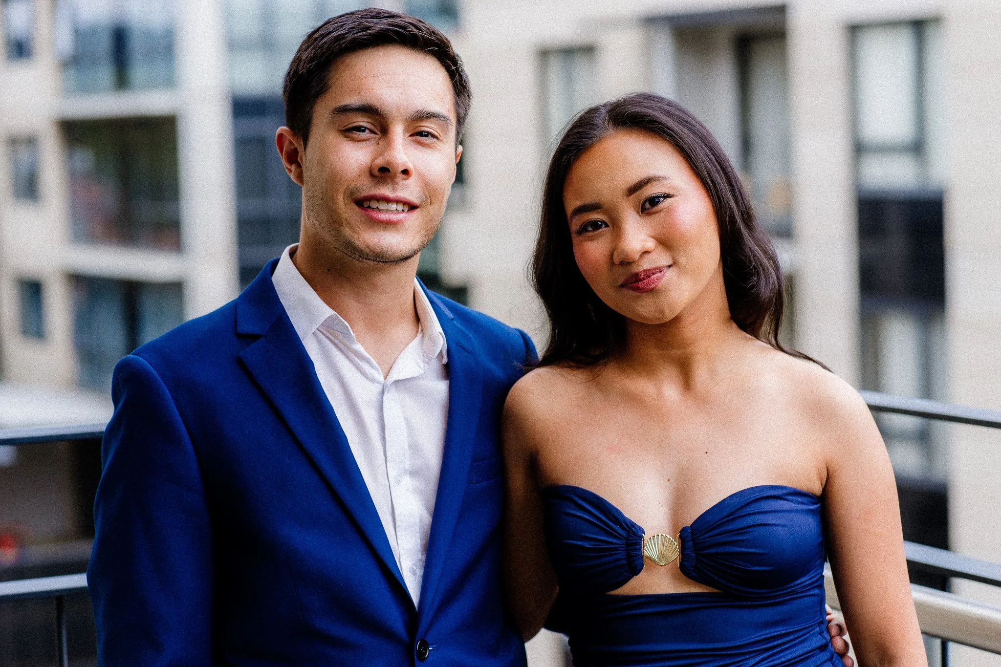 A man in a blue suit and white shirt standing next to a woman in a strapless blue dress, both smiling outdoors with a building in the background.