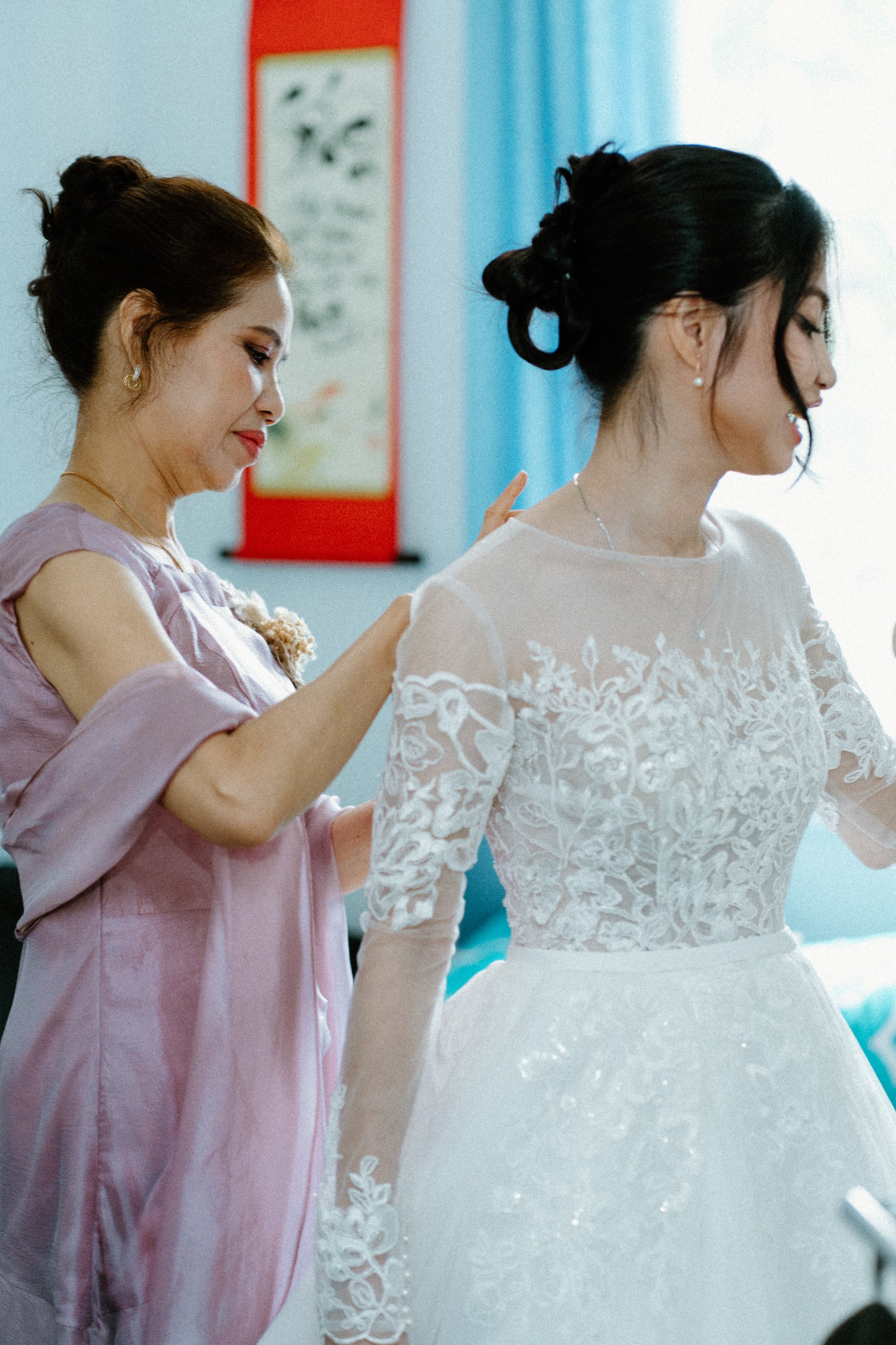 A woman in a white lace wedding dress stands with her back to the camera, while another woman in a pink satin dress helps her with the final touches. The room has a blue curtain and a red and white decorative panel in the background.
