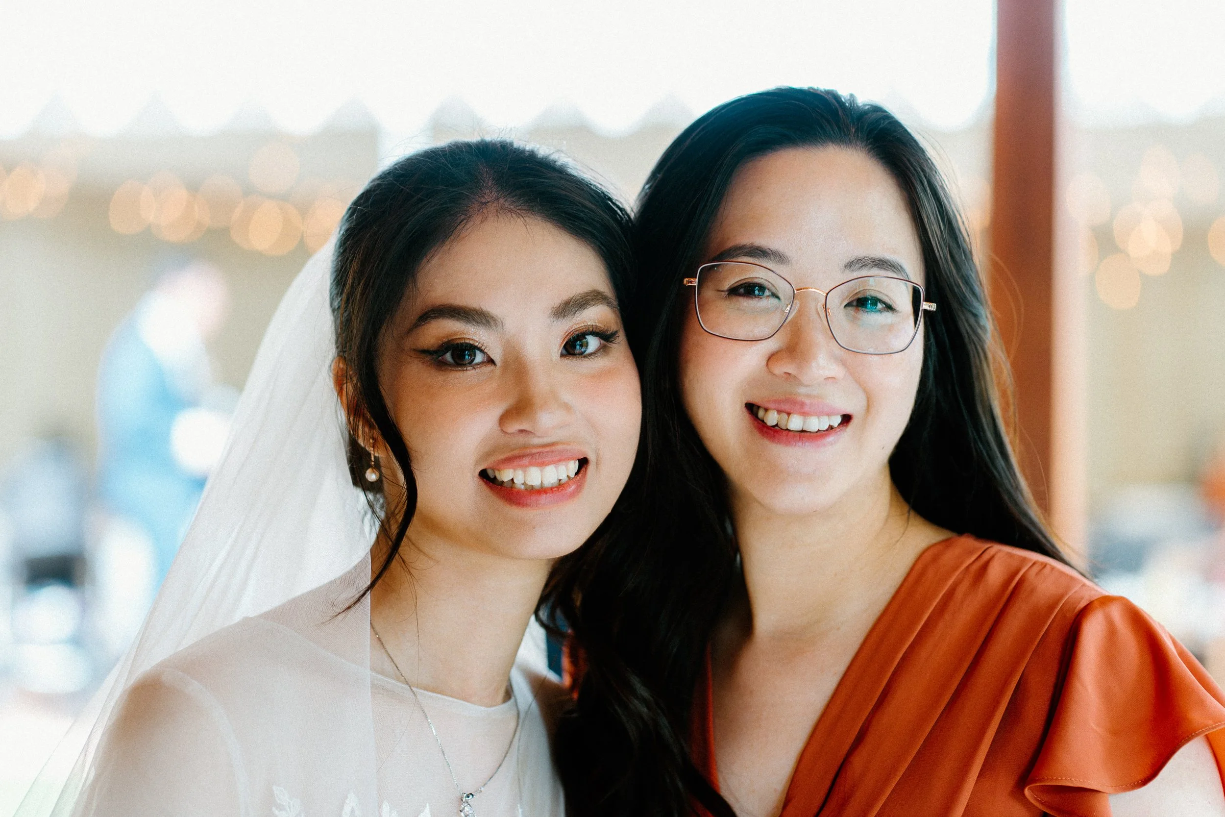 Two women smiling, one wearing a wedding dress and veil, the other in an orange dress and glasses, standing close together at a celebration.