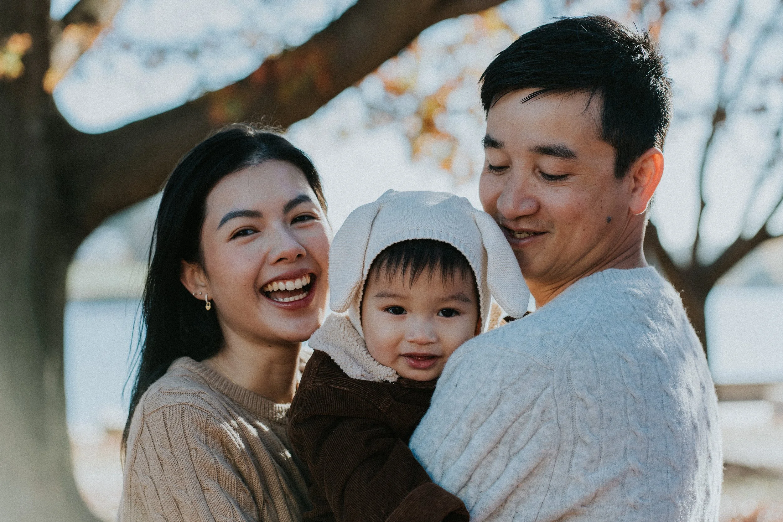 A happy Asian family of three, including a woman, a man, and a young child, outdoor with trees in the background during daytime.