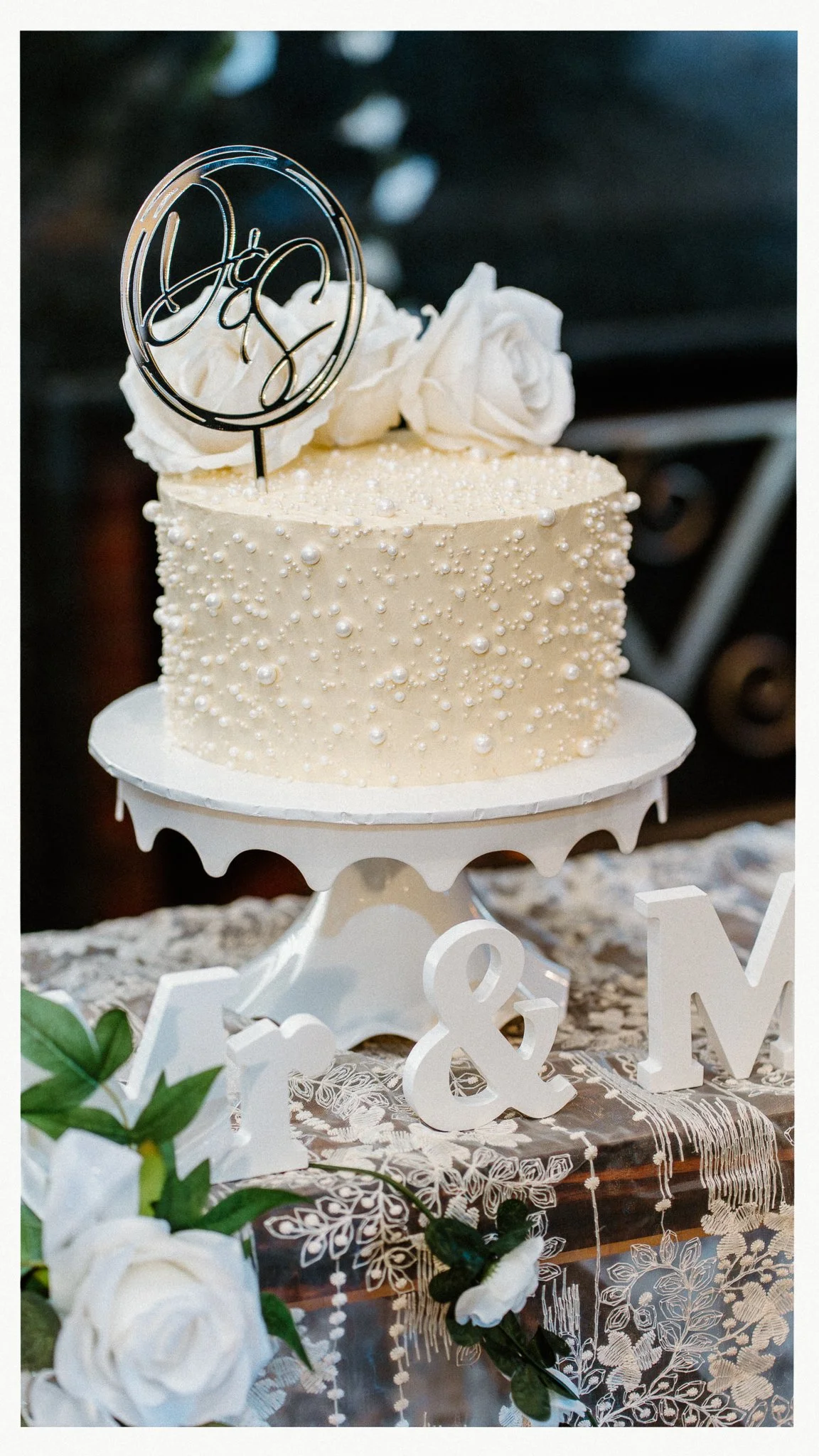 White wedding cake decorated with small pearls and white roses, topped with a black 'Best' cake topper, displayed on a white stand with decorative accents, surrounded by white roses and greenery.