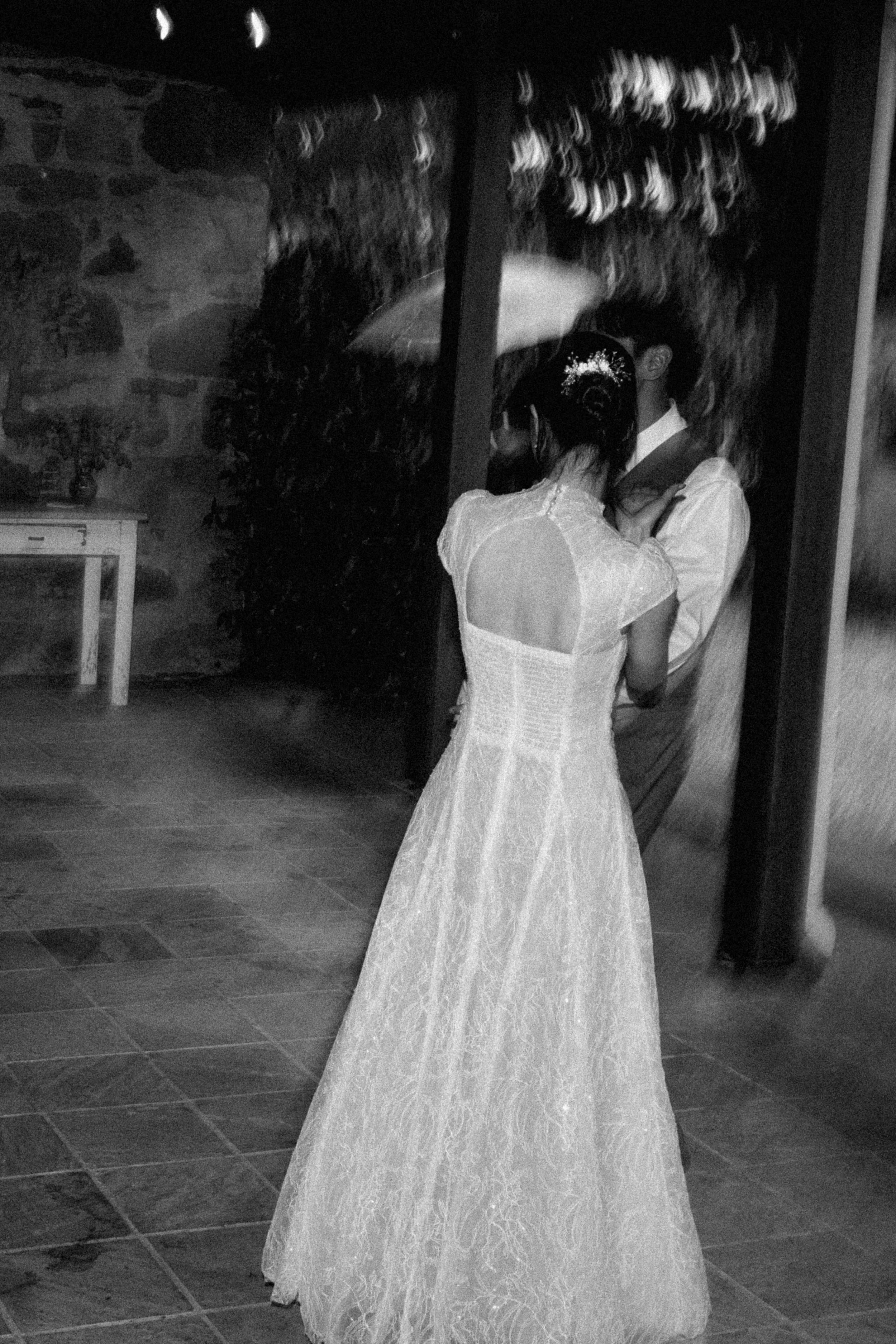 A couple sharing a kiss in a wedding dress and suit, standing outdoors at night, seen through a glass reflection.