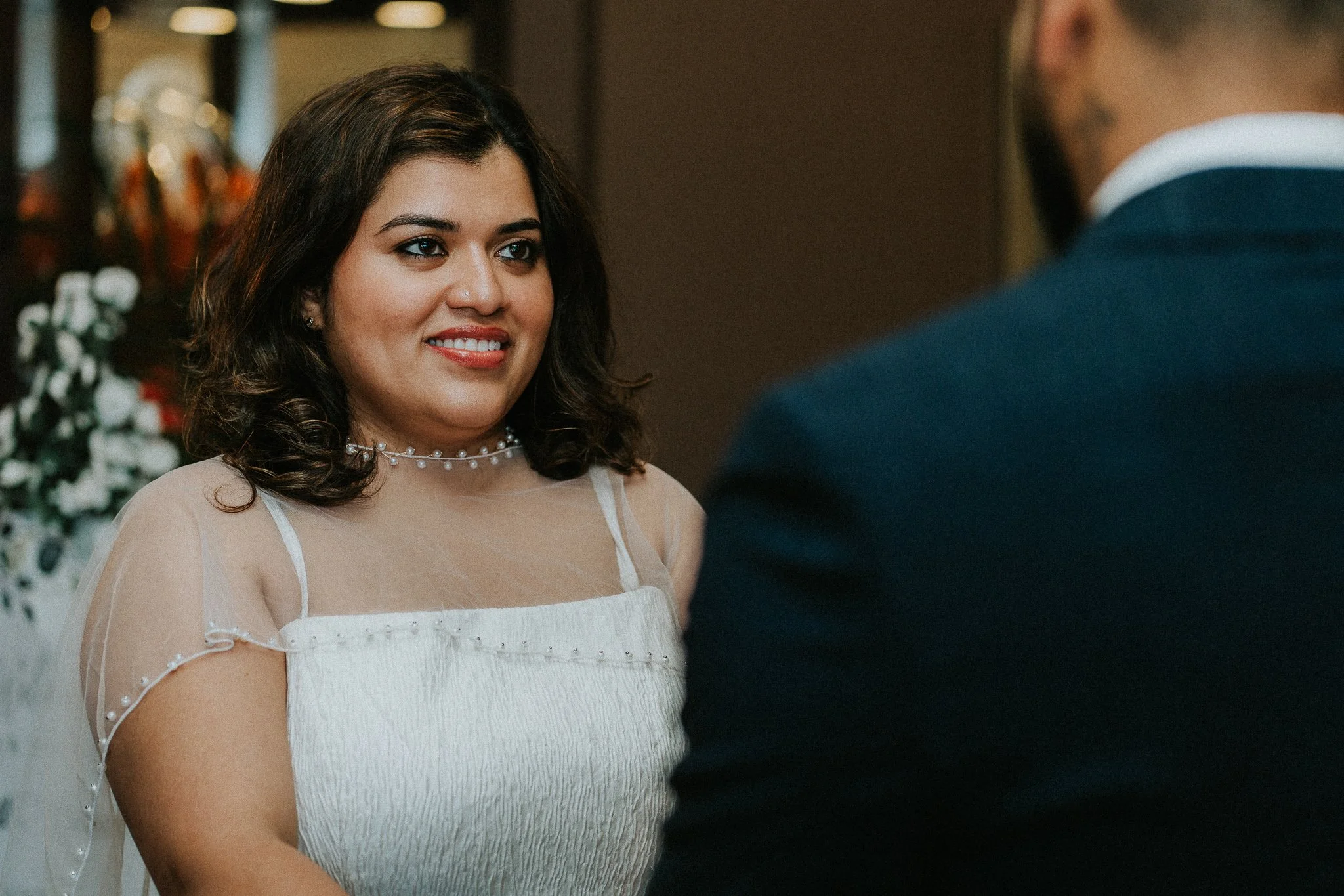 A woman with dark brown, curly hair, wearing a white dress with sheer, short puffed sleeves and pearl embellishments, smiling at a man in a dark suit during a wedding or formal event.