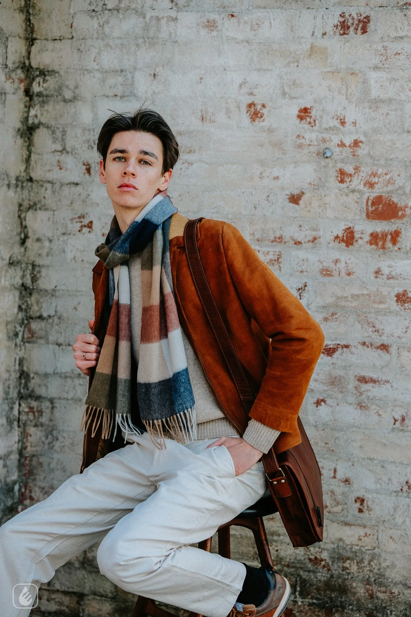 A young man sitting on a wooden chair against a weathered brick wall. He is wearing a tan and beige sweater, a multicolored plaid scarf, white pants, and brown shoes. He has dark hair and a serious expression, with a brown leather bag resting on the floor beside him.