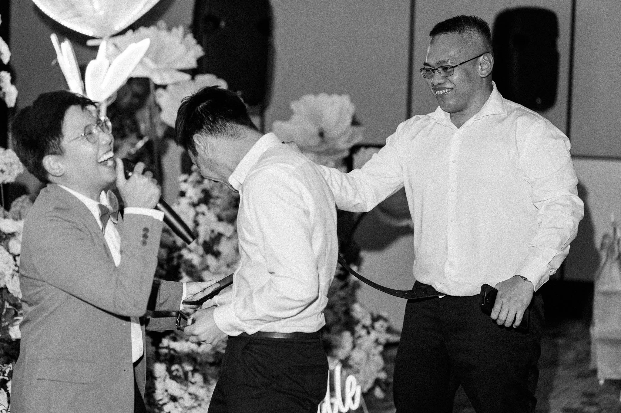 Three men engaging in a handshake or greeting, with a background featuring flowers and decorative elements, in a celebration or event setting.