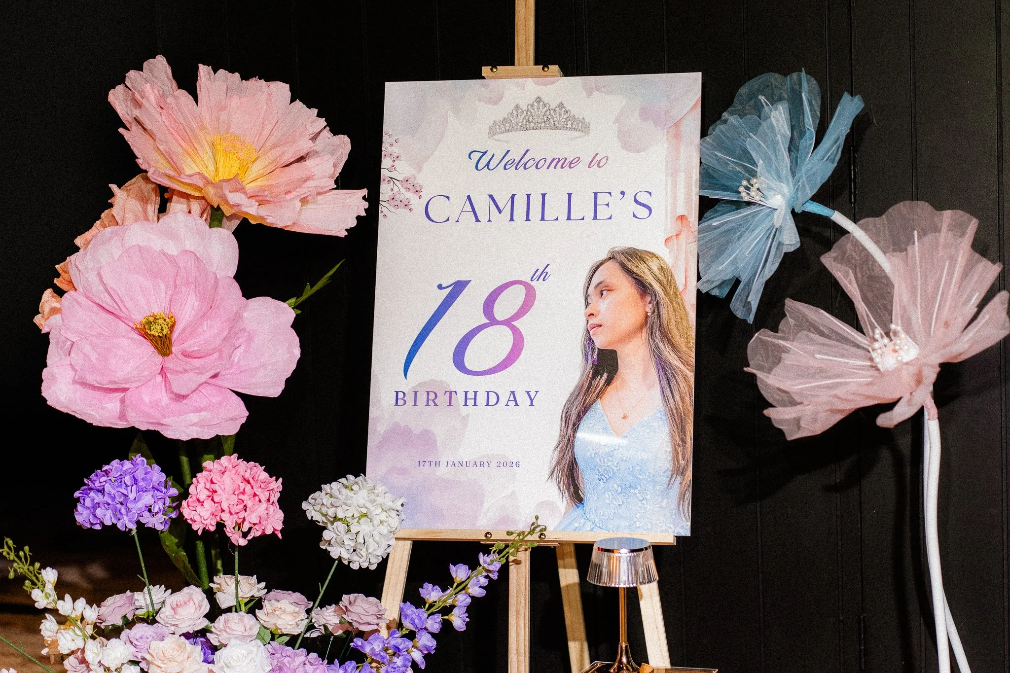 A birthday celebration setup with a sign that reads "Welcome to Camille's 18th Birthday" dated January 17th, 2026. The décor includes large pink, purple, and white paper flowers arranged around the sign, which features a crown graphic and a photo of 