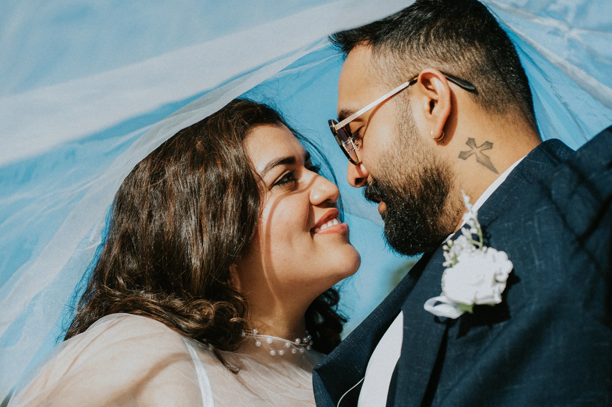 Close-up of a smiling couple at their wedding with their foreheads touching under a sheer veil against a clear blue sky.