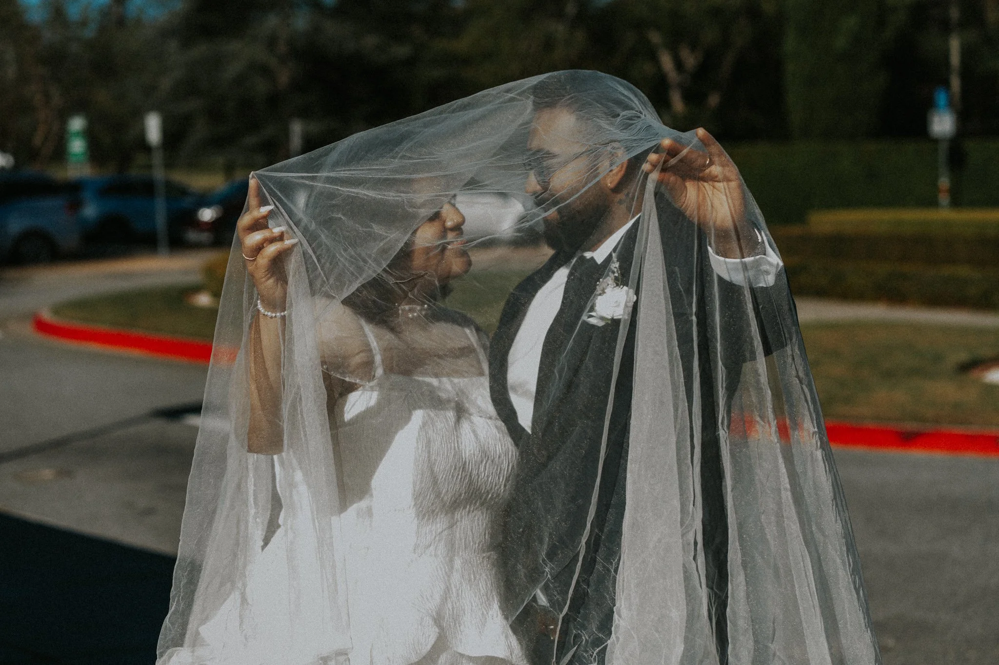 A bride and groom standing close together outside, celebrating their wedding, with the bride holding a sheer veil over both of their faces.