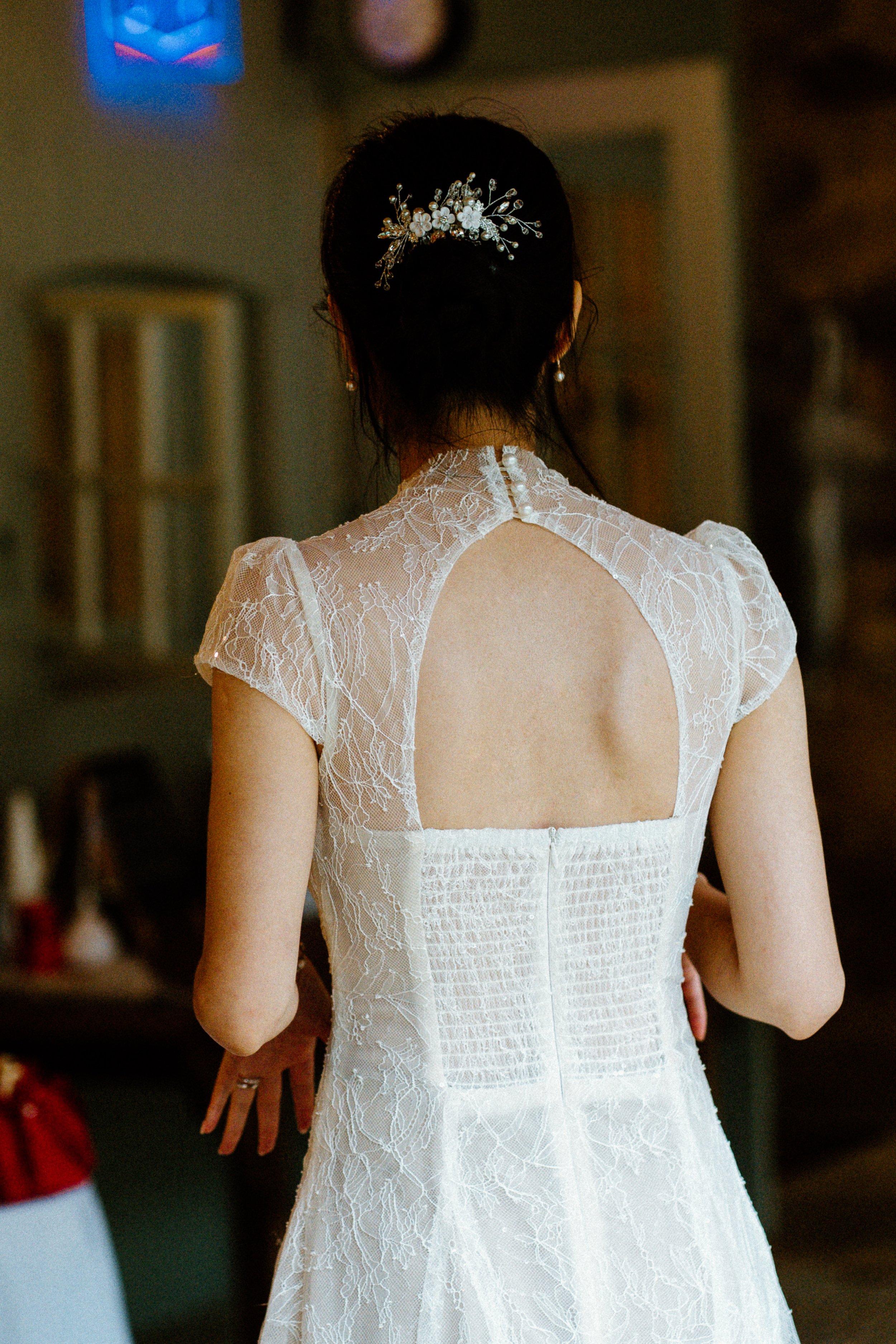 Back view of a woman wearing a white lace wedding dress with an open back, adorned with a floral hairpiece, in a room with dim lighting.