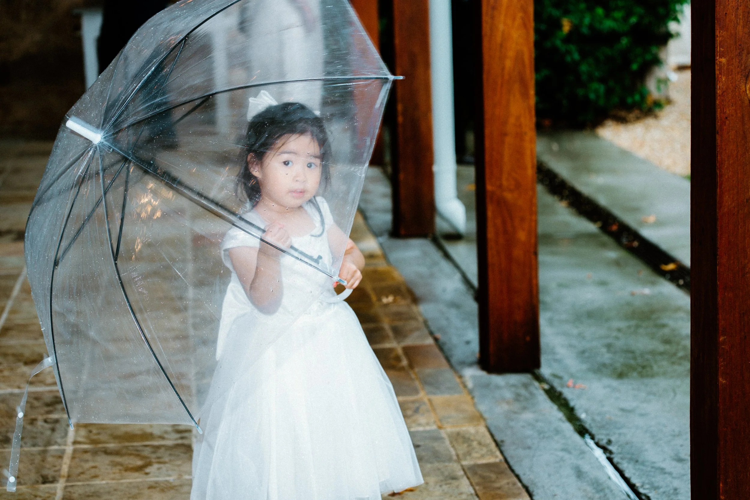 A young girl in a white dress holding a transparent umbrella, standing on a wet outdoor walkway next to a wooden railing on a rainy day.