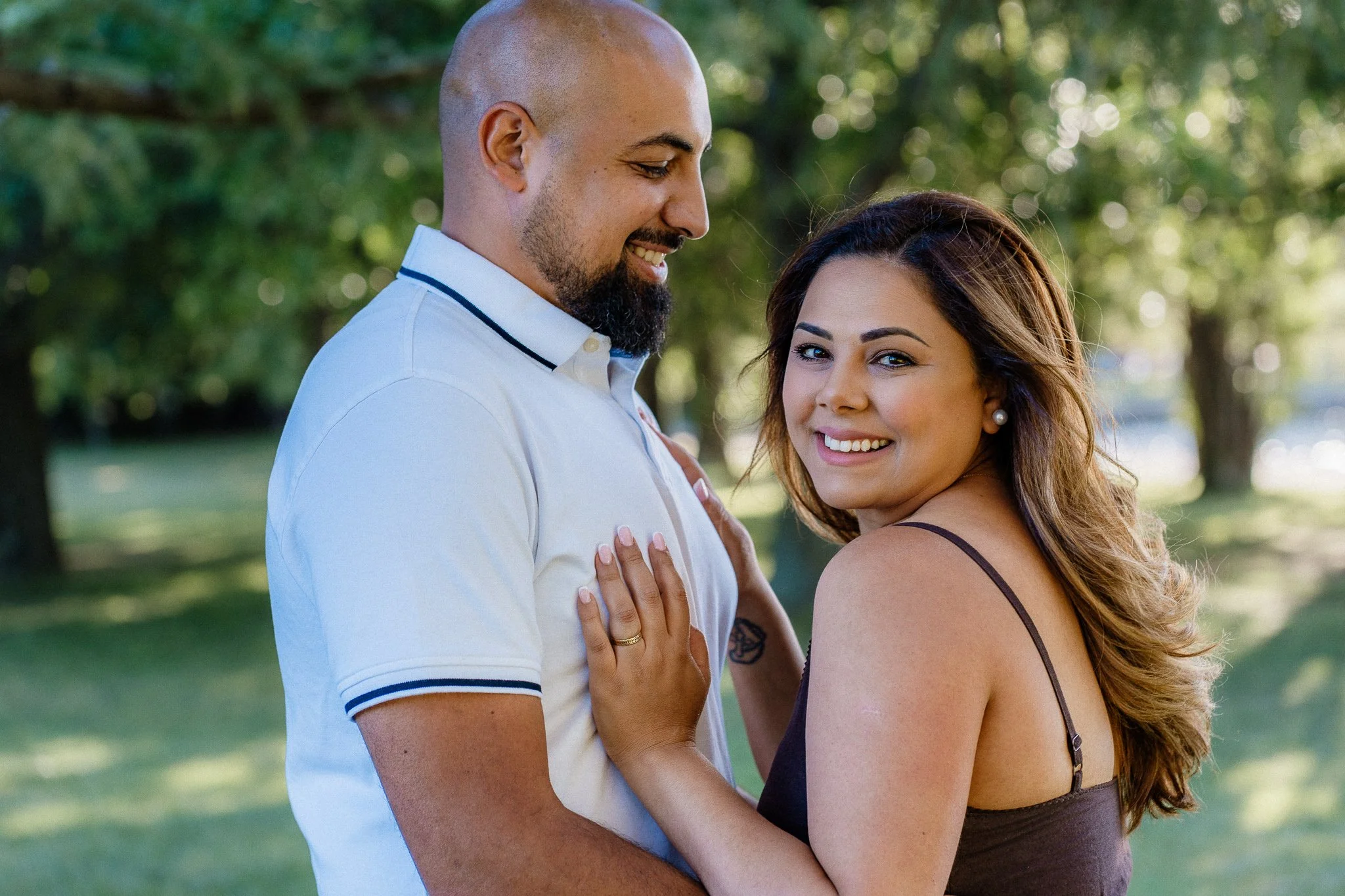 A couple smiling and embracing outdoors in a park with green trees in the background. The man is bald with a beard, wearing a white polo shirt. The woman has long brown hair, wearing a dark strappy top and pearl earrings.