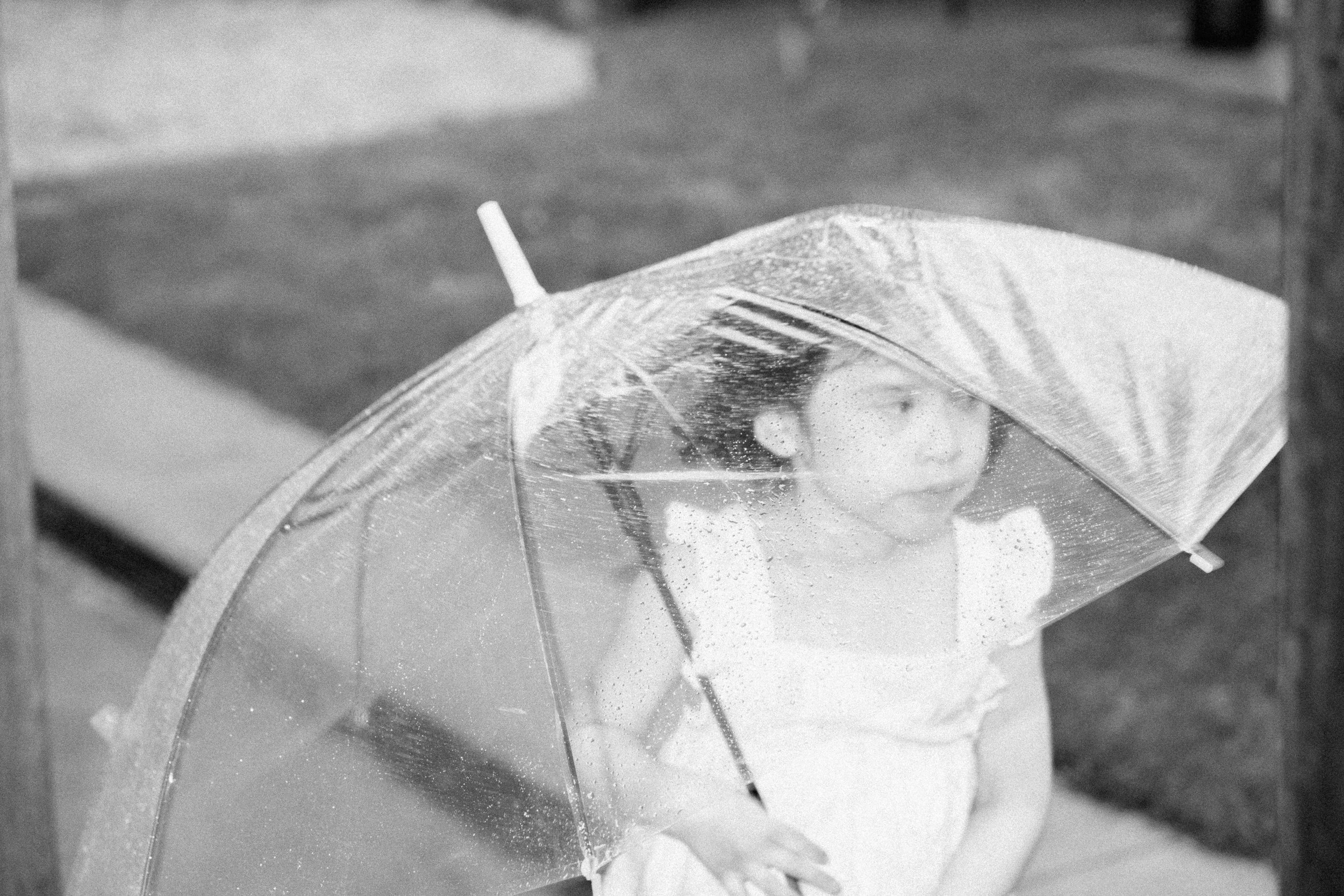 A woman with glasses sitting inside a bubble-shaped transparent umbrella during a rainy day.