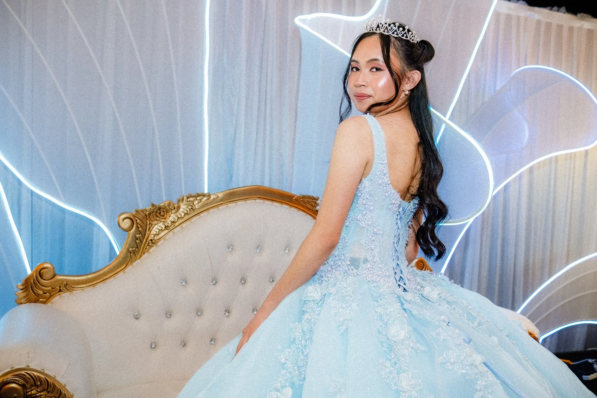 A young woman wearing a light blue dress and a tiara sitting on an ornate chair in front of a neon light backdrop.