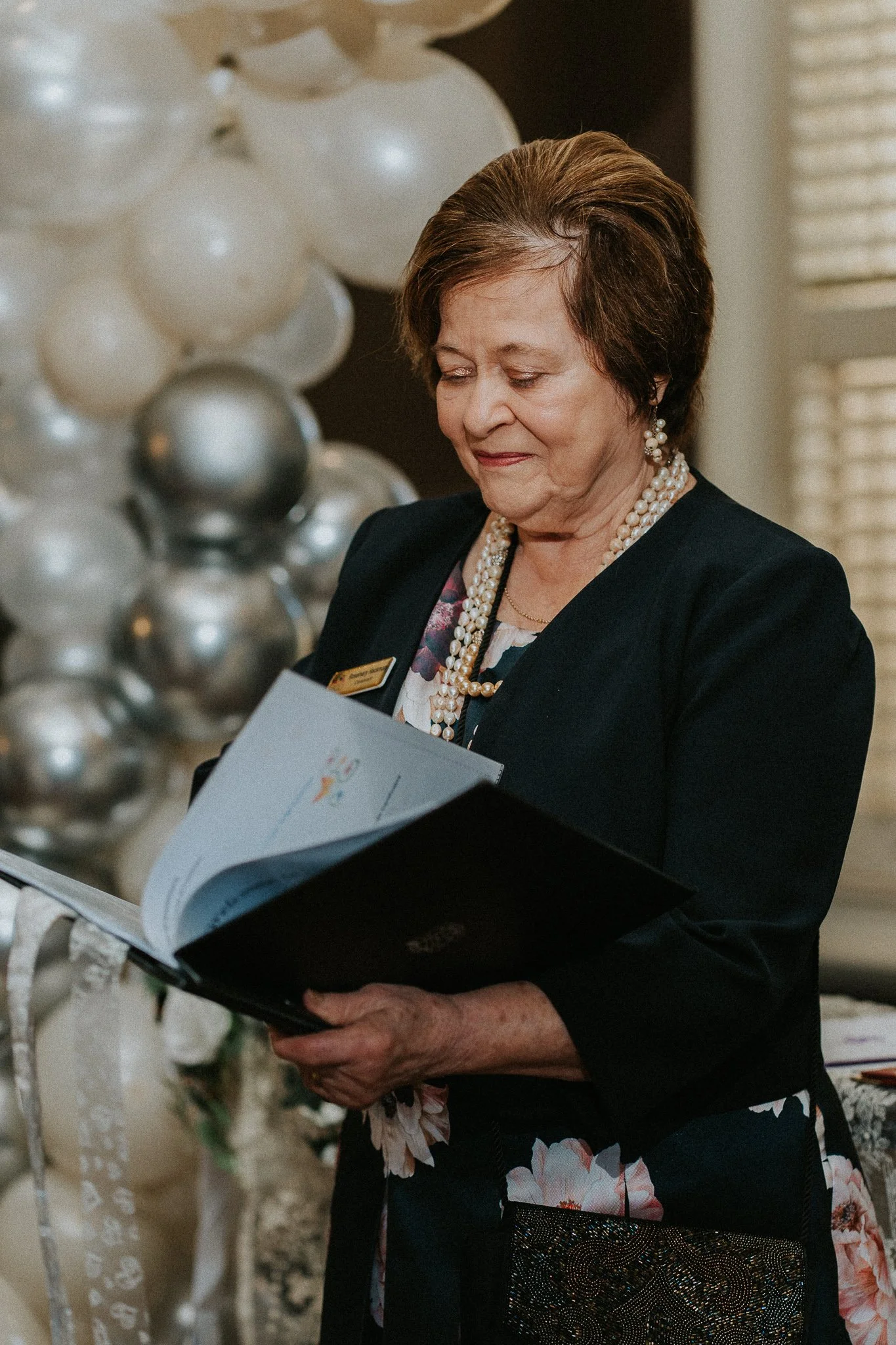 An older woman with short brown hair, wearing pearl earrings, a pearl necklace, and a floral dress, is looking at a booklet while standing at a formal event with a balloon decoration in the background.