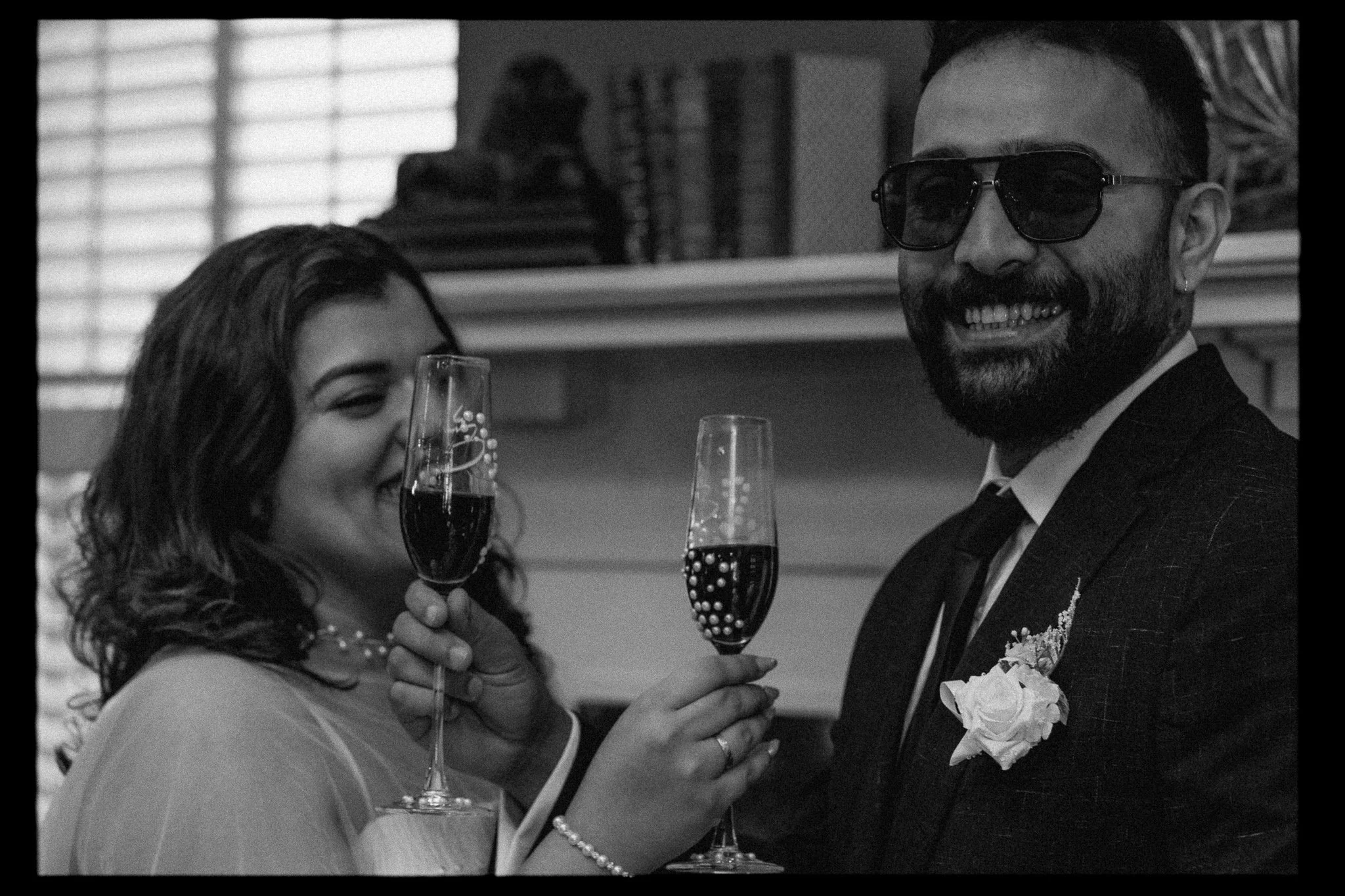 A woman and a man are smiling and holding glasses of champagne, celebrating together. The woman is wearing a white dress and a pearl bracelet, while the man is dressed in a tuxedo with a boutonniere and sunglasses.