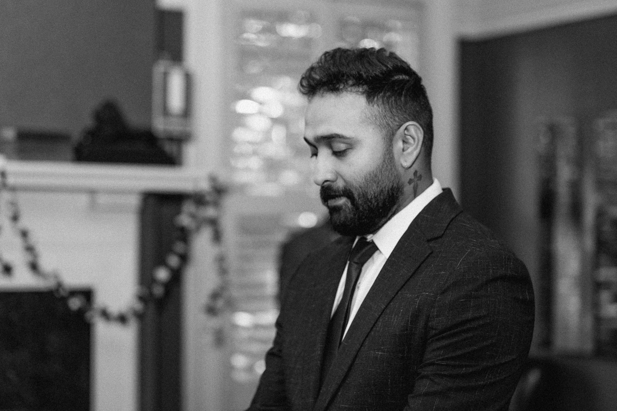 A man in a suit with closed eyes, a beard, and a tattoo on his neck, standing indoors in a room with blurred background.
