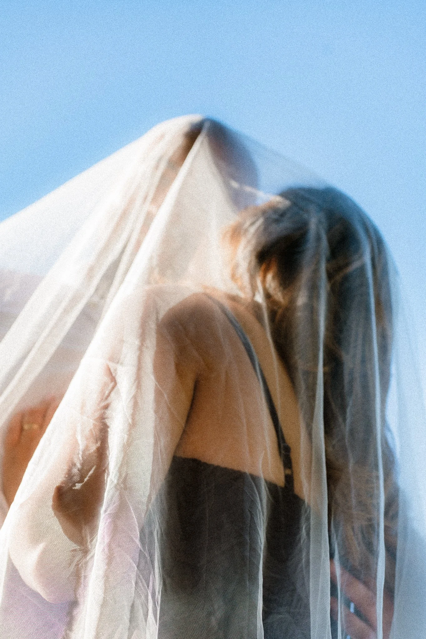 A woman with long hair wearing a dress and a black strap top, standing behind a sheer curtain against a blue sky.