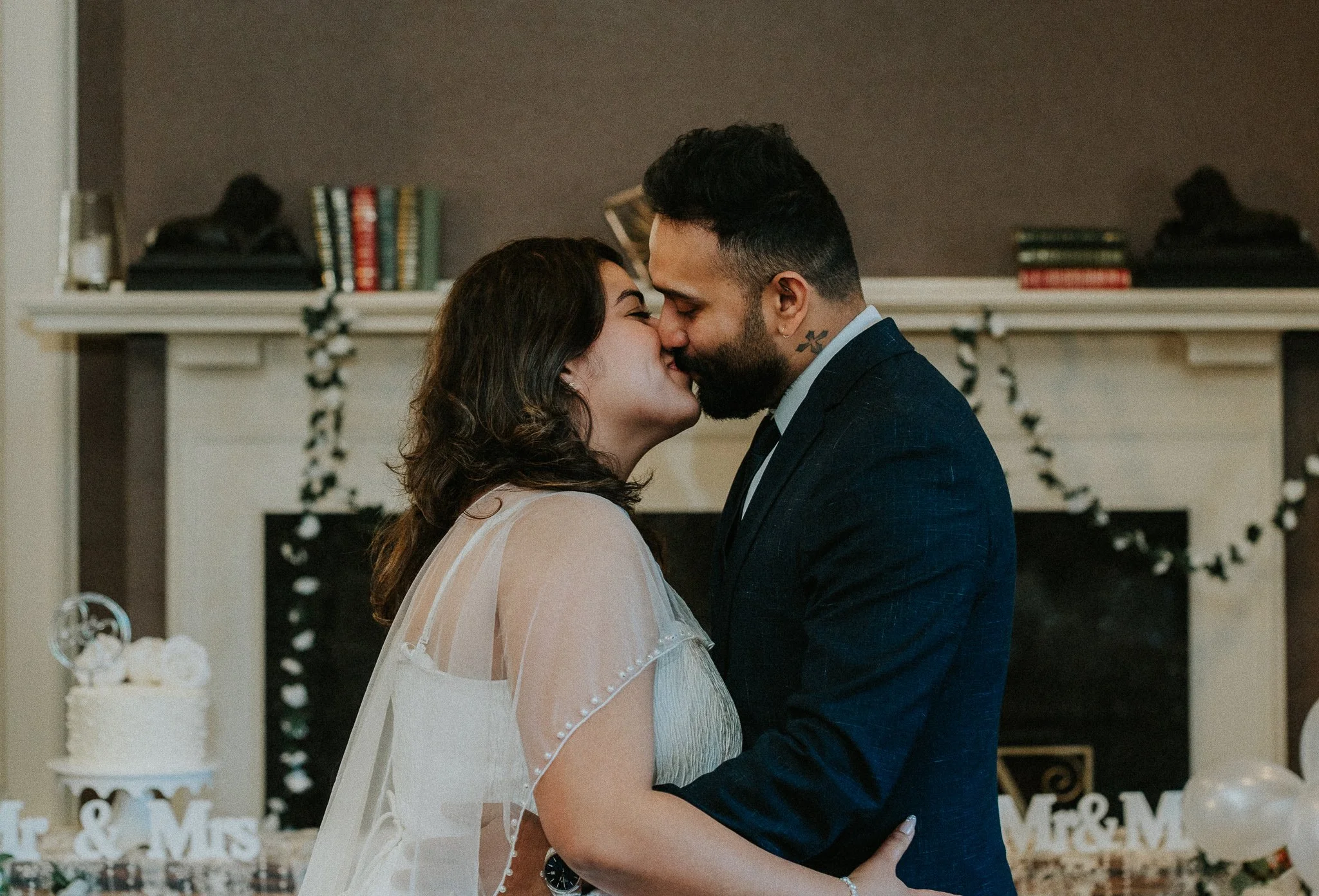 A couple kisses during their wedding ceremony in front of a decorated fireplace mantel with books and holiday decorations.
