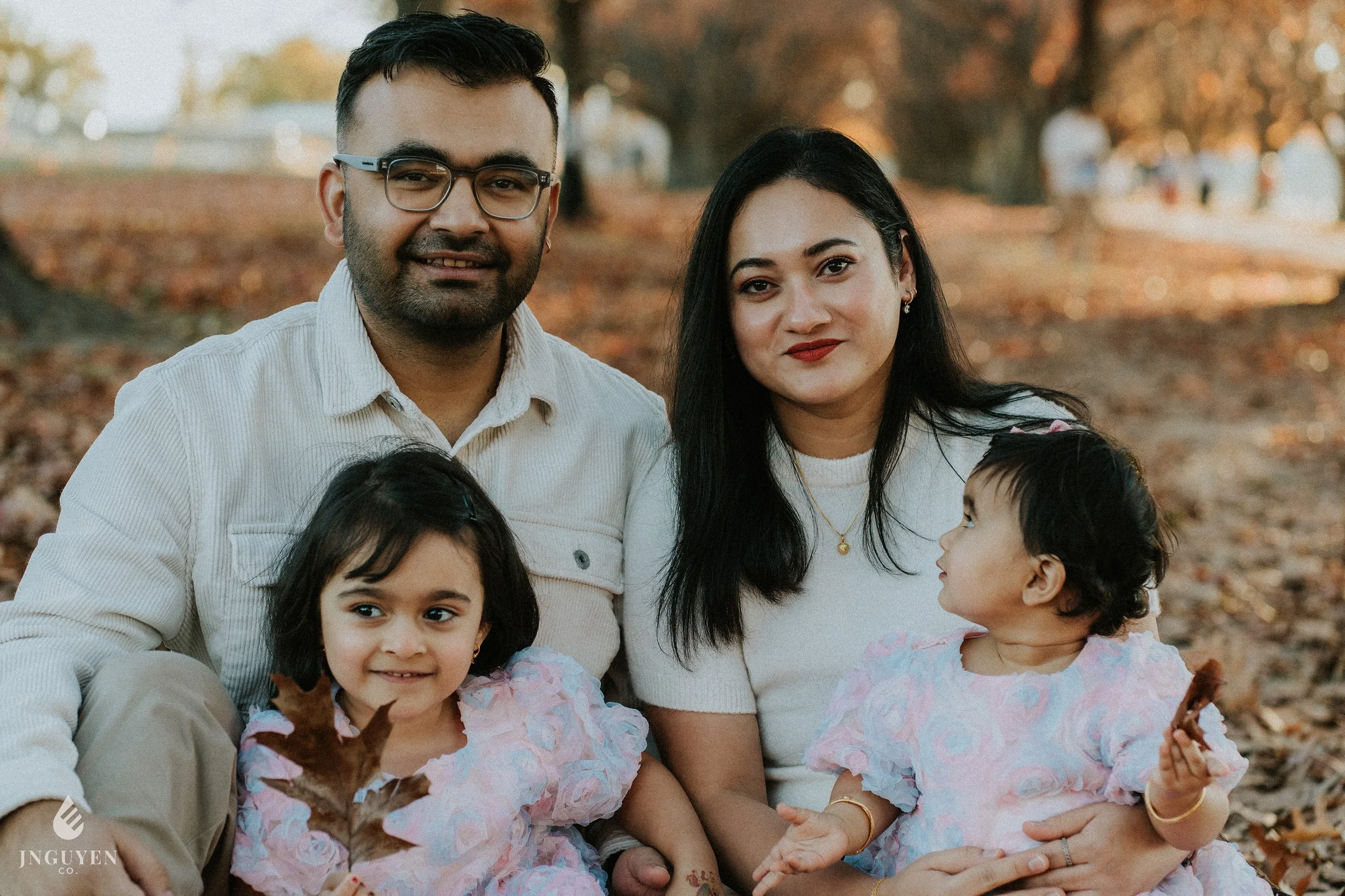 A multi-generational family outdoor portrait during fall, including a man with glasses, a woman, and three young children, all smiling and sitting on autumn leaves.