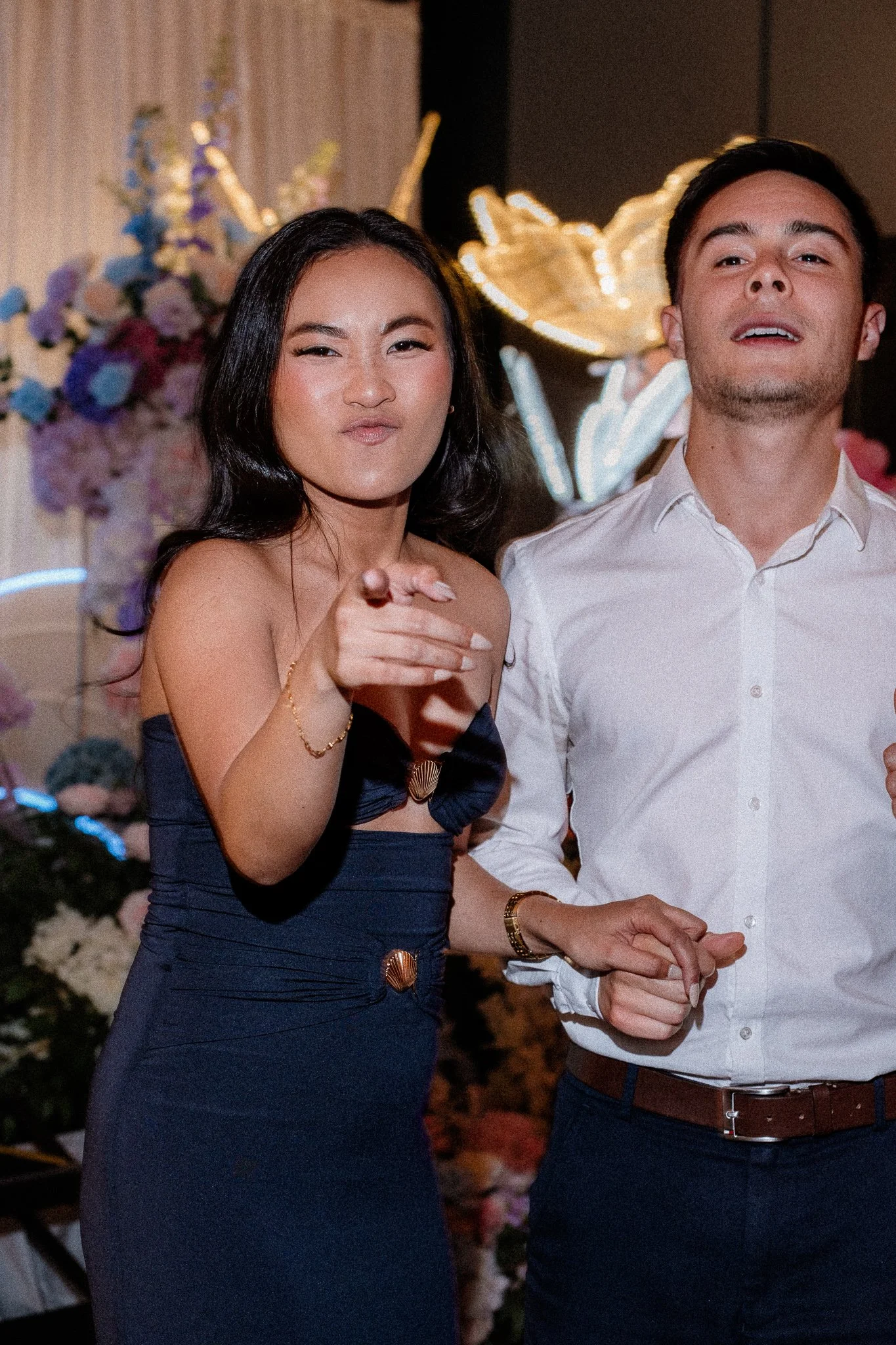A woman in a dark blue dress with a gold bracelet and necklace is pointing her finger at the camera. She has long dark hair. Next to her, a man in a white shirt with rolled-up sleeves and a brown belt is standing with his hands clasped. The backgroun