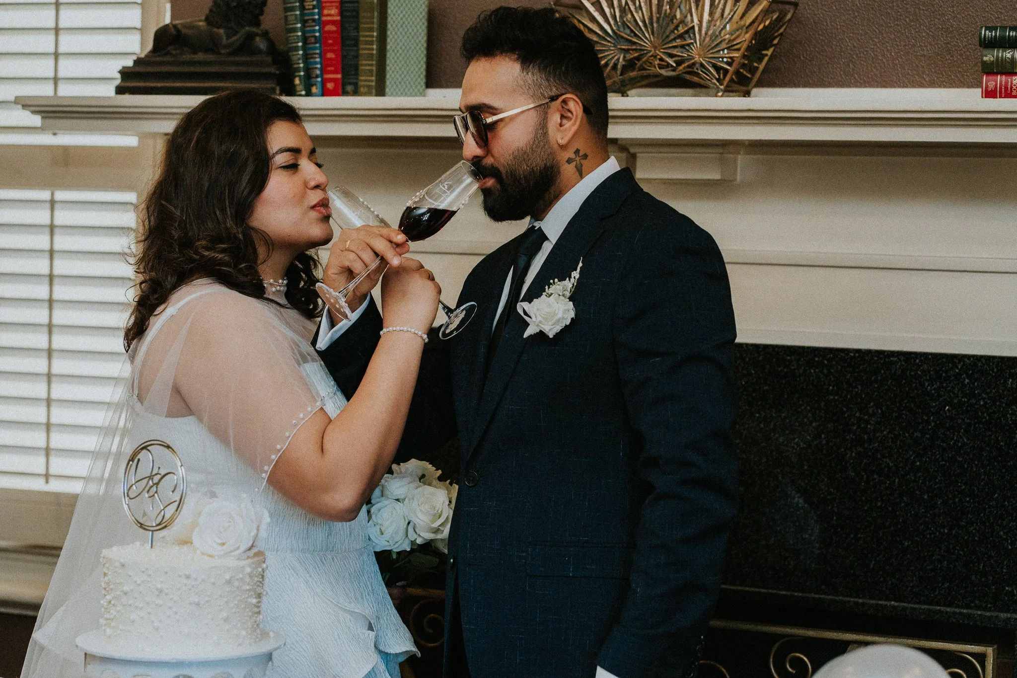 A bride and groom sharing a toast with red wine at their wedding reception. The bride is dressed in white with dark hair and the groom in a suit with glasses and a tattoo on his neck. The cake with a topper is visible in the foreground.