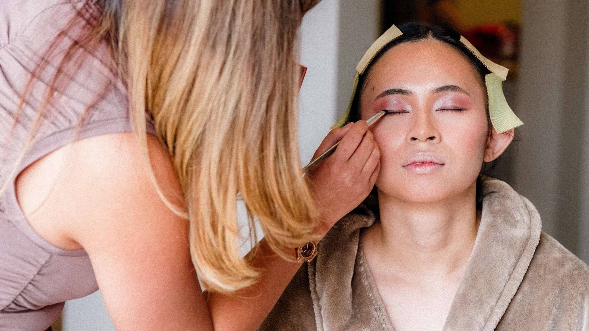 Makeup artist applying eyeshadow to a woman's closed eyes, with hair rollers and a hoodie, in a close-up shot.