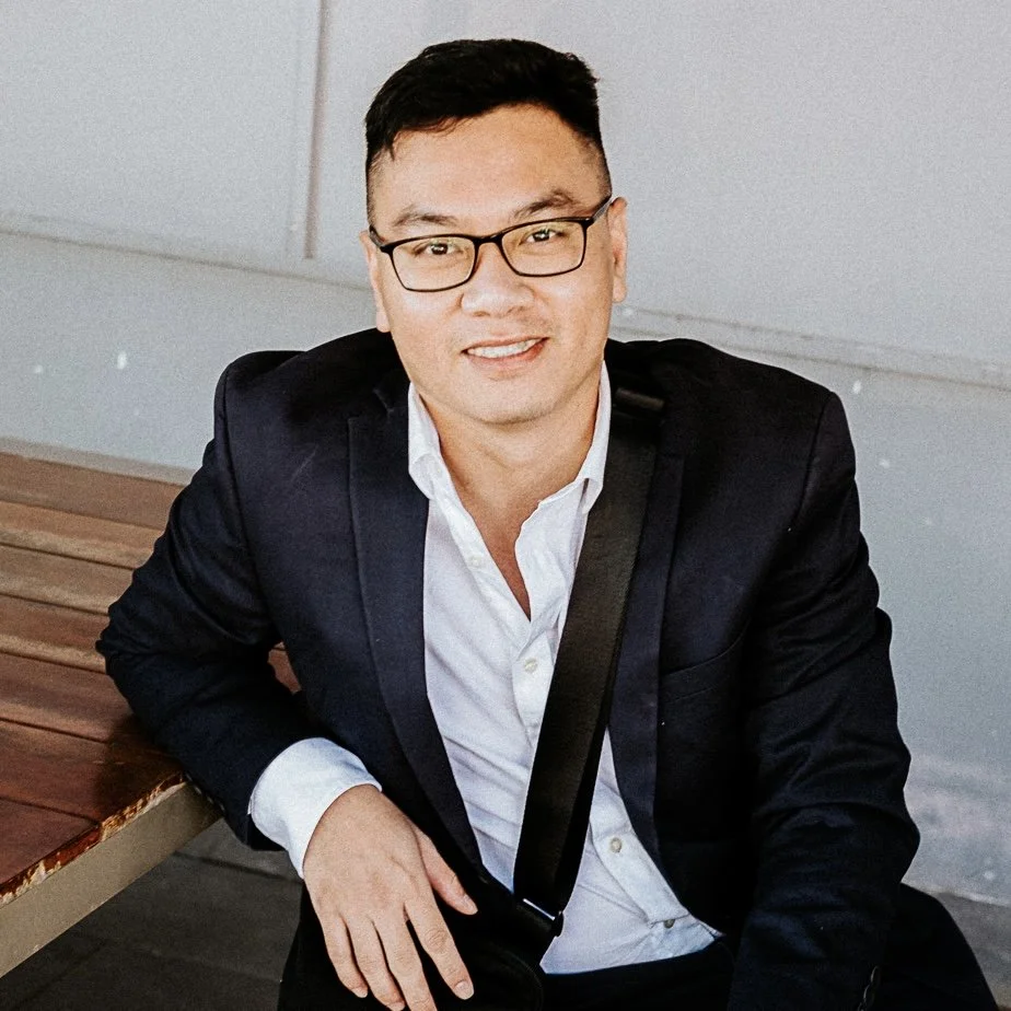 A man in glasses wearing a black suit and white shirt sitting at a wooden table.