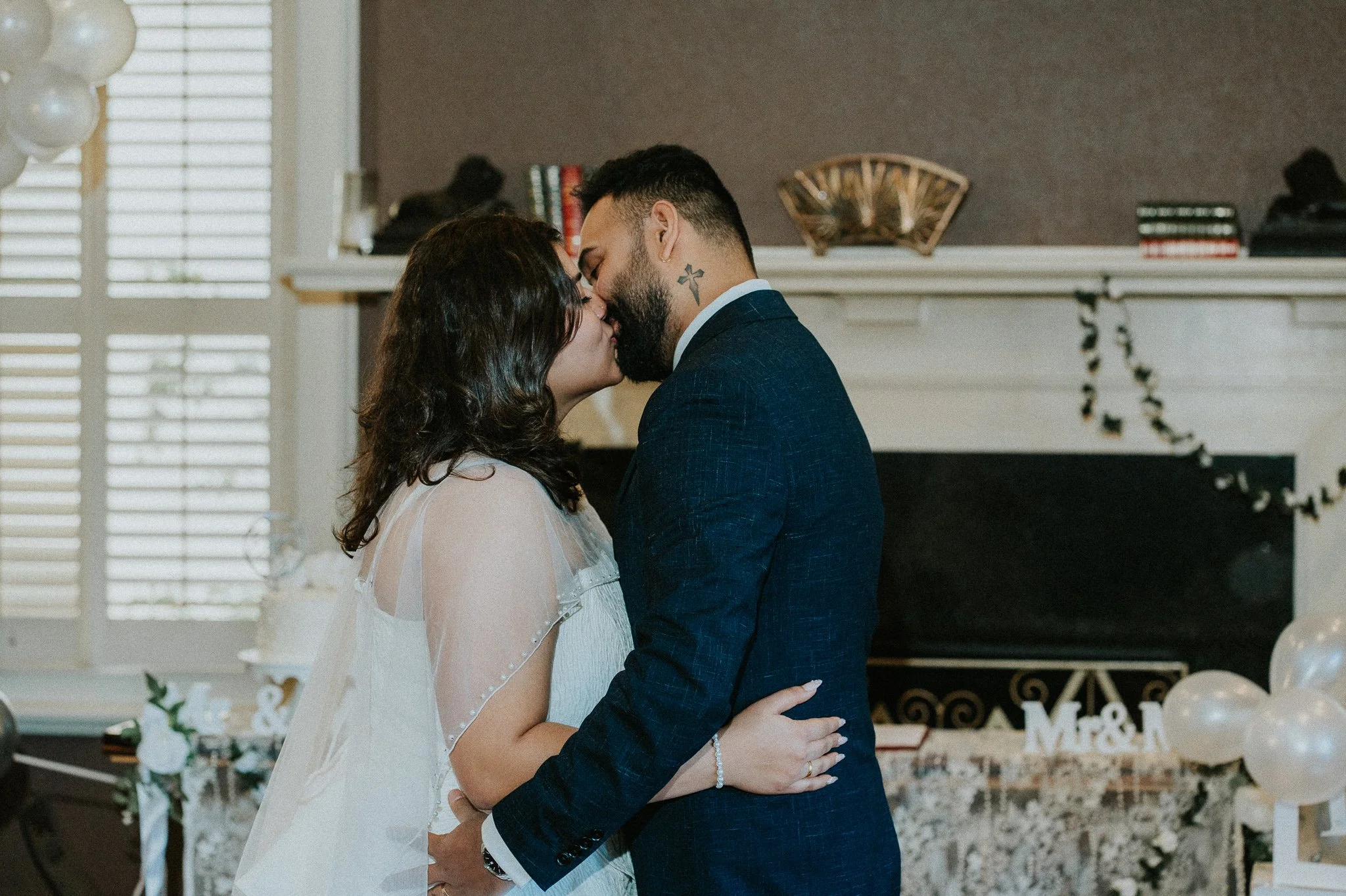 A bride and groom embrace and prepare to kiss during their wedding in a decorated indoor setting, with white balloons, flowers, and a sign that says 'Mr. & Mrs.' in the background.