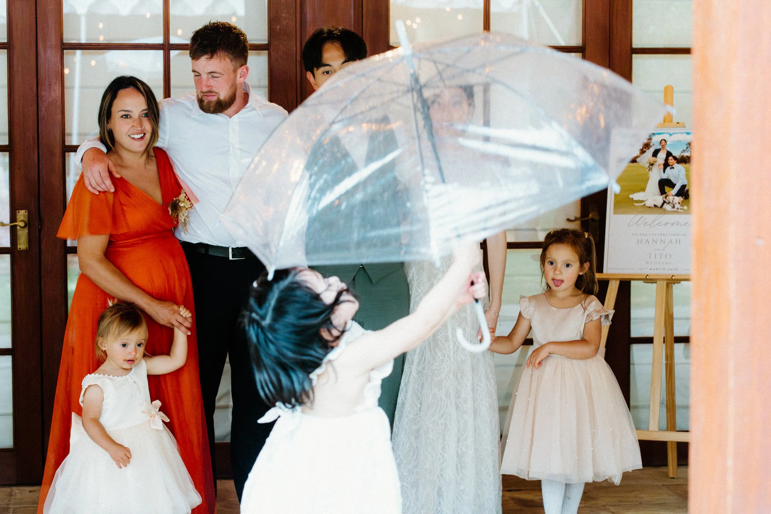 A group of children and adults at a wedding reception in a decorated indoor venue. A woman in a white dress, likely the bride, is holding a clear umbrella. Two young girls in white dresses and an older woman in a red dress are visible, with the other