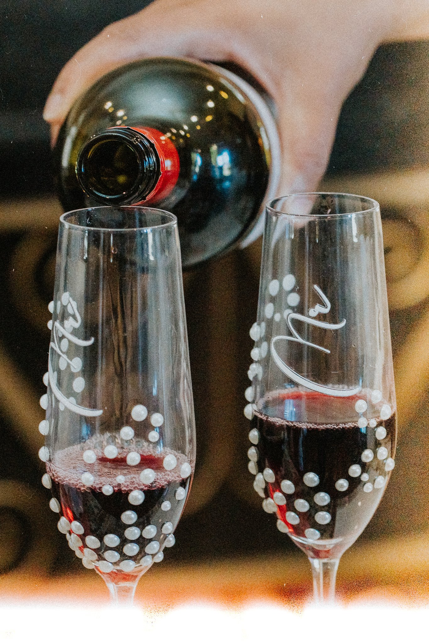 Two glasses of red wine on a table, with a person pouring another glass of wine in the background. The glasses are clear with white polka dot patterns.