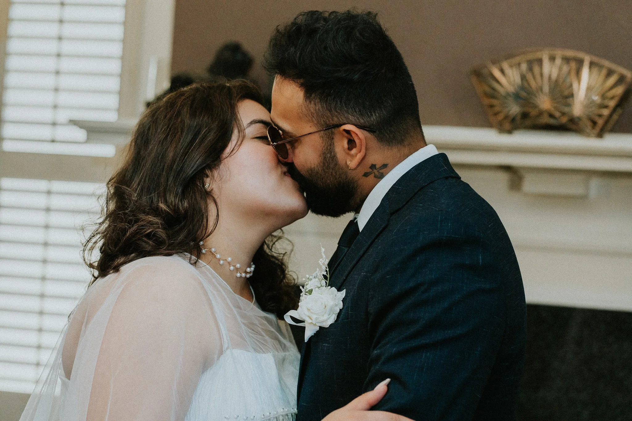 A bride and groom share a kiss during their wedding ceremony, with the bride wearing a white dress and pearl necklace, and the groom in a dark suit with sunglasses and a tattoo on his neck.