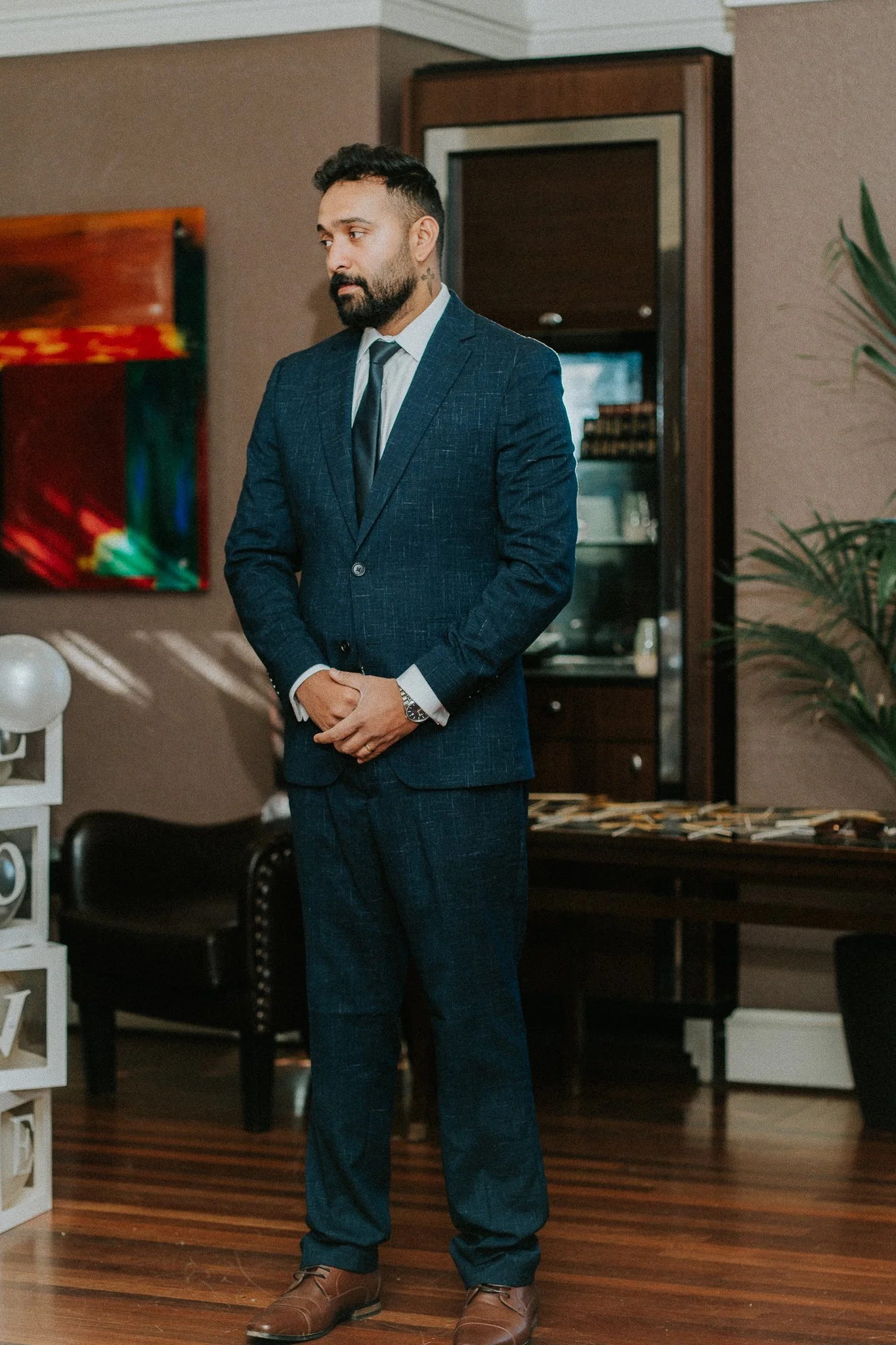 A man in a dark blue suit, white shirt, and black tie stands indoors with his hands clasped in front of him. He has a beard, short dark hair, and is wearing brown shoes. The background includes a colorful abstract painting, some furniture, and a beig