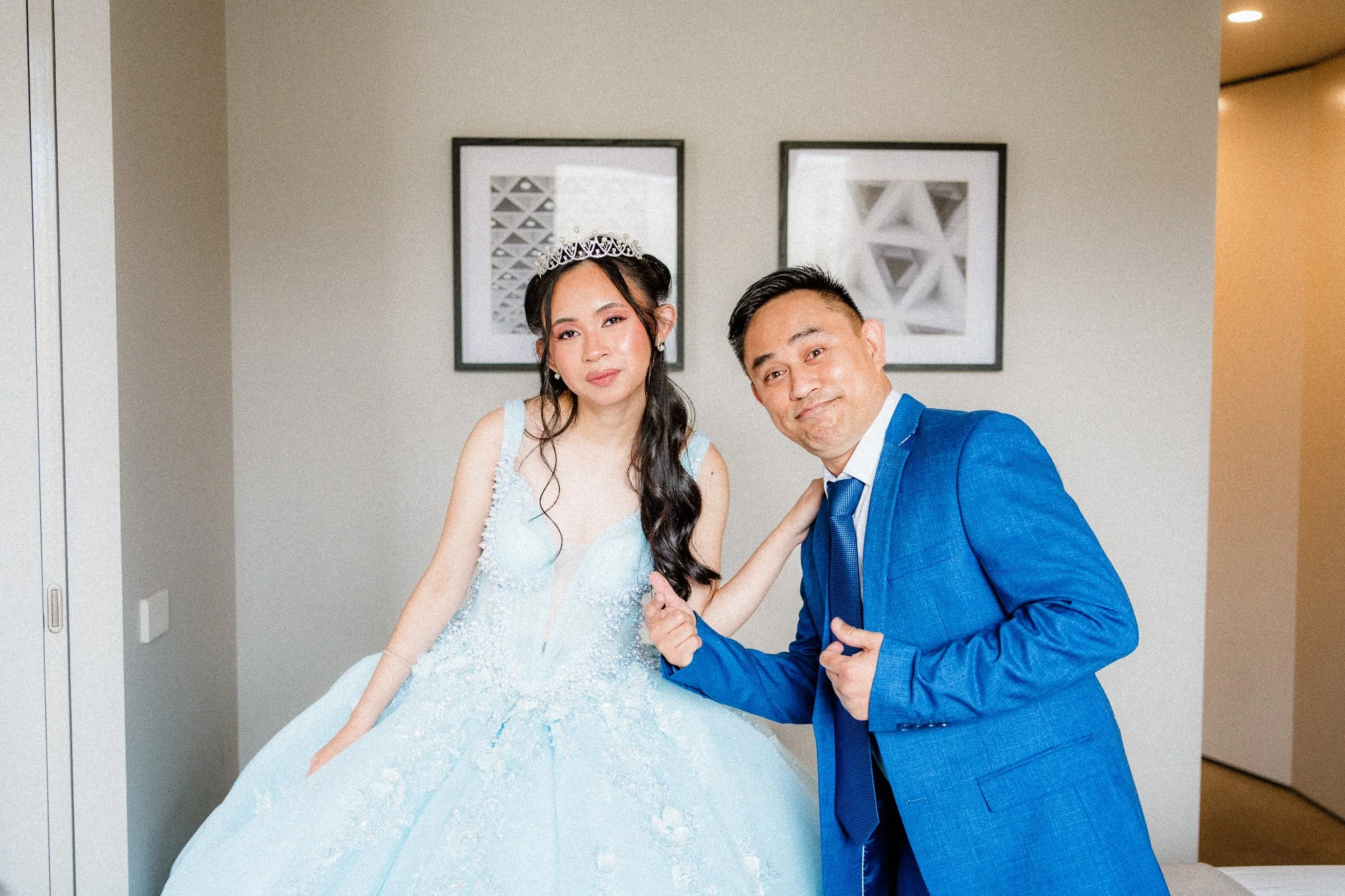 A young woman in a light blue, beaded dress with a tiara, and a man in a blue suit pose together indoors with framed abstract geometric art on the wall behind them.