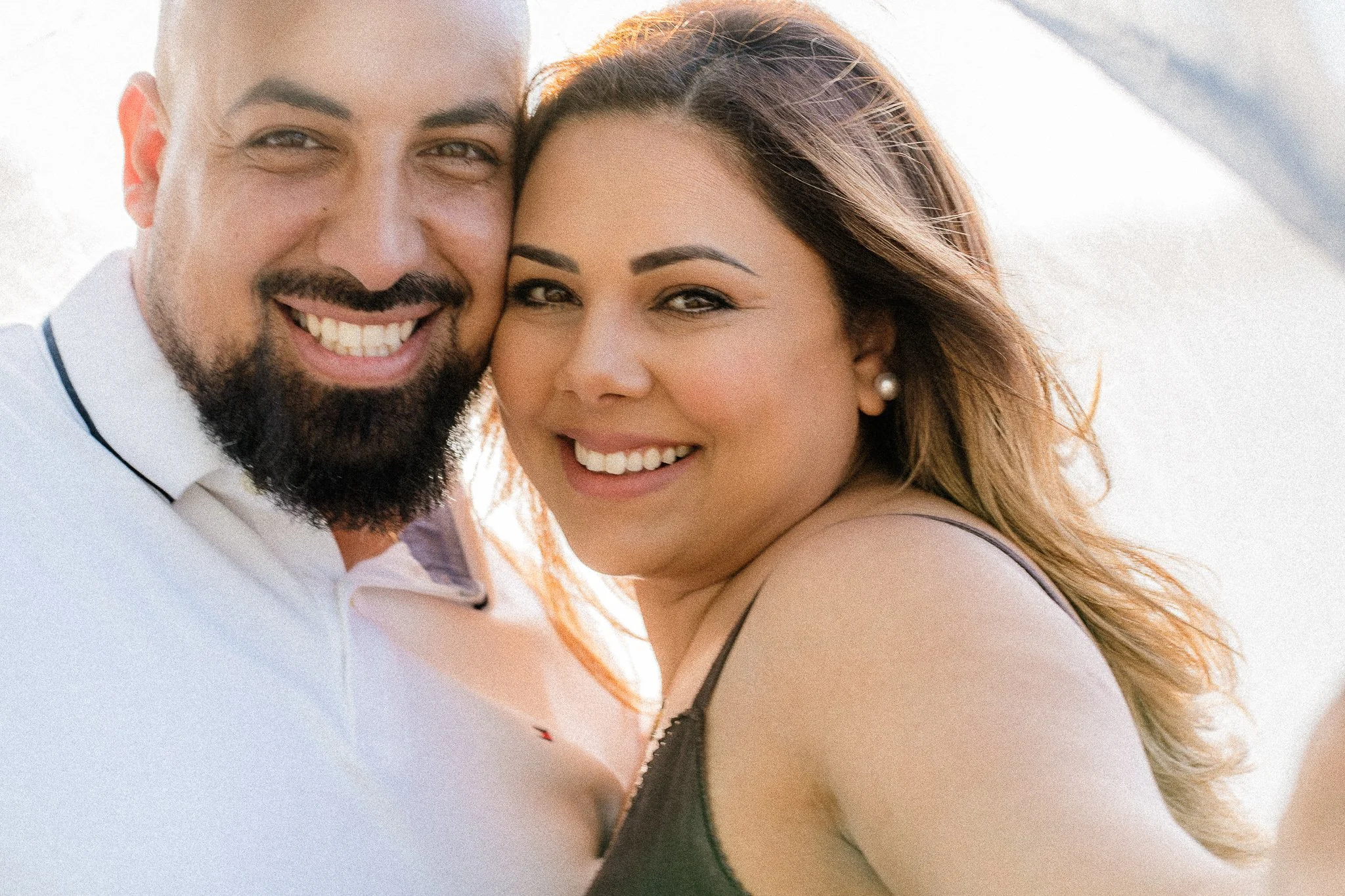 A happy couple smiling, with the man having a beard and the woman wearing pearl earrings, close-up shot outdoors.