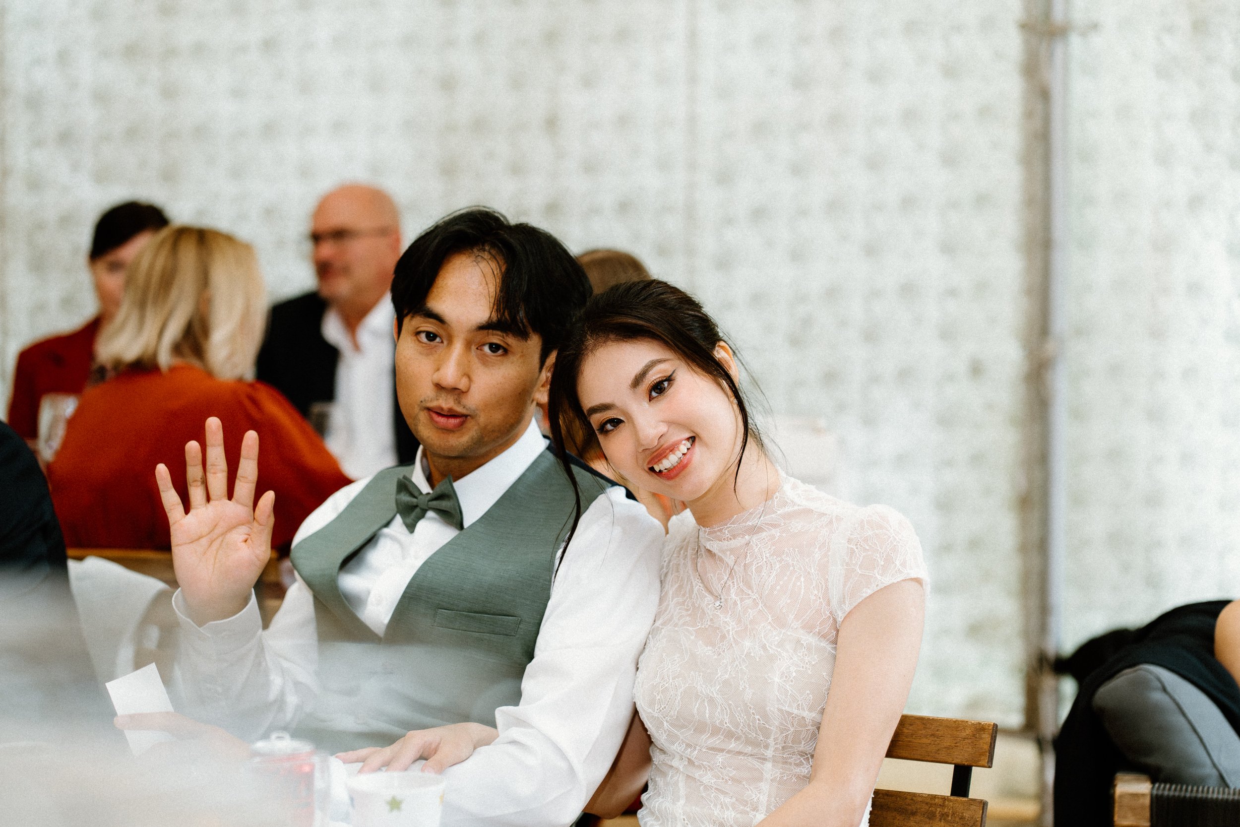 A young woman and man sitting at a table, smiling and waving at the camera, with several other people in the background.