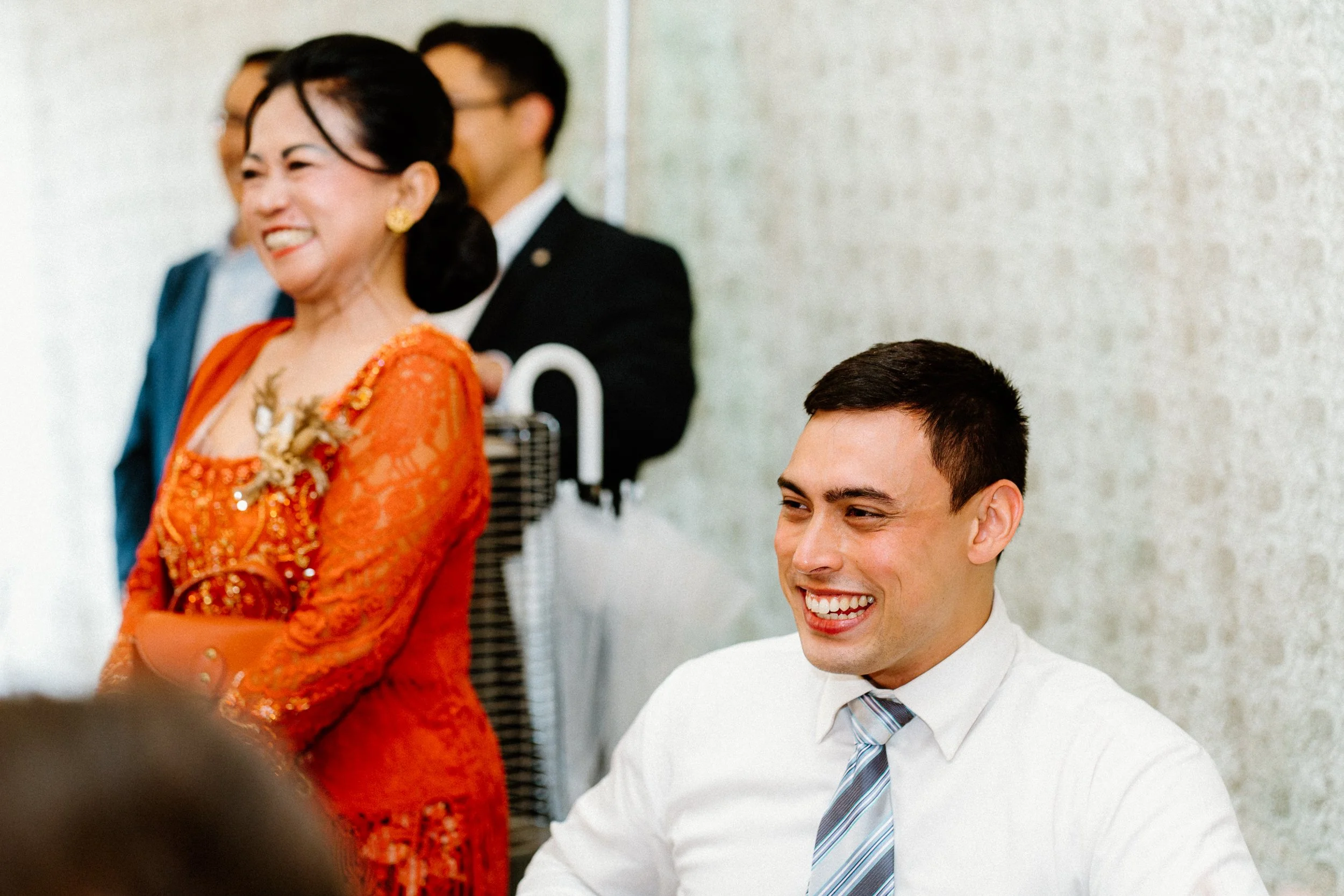 A group of people in a professional setting, with a woman dressed in traditional orange attire smiling and standing, and a man in a white shirt and striped tie seated and smiling in the foreground. Others are partially visible in the background.
