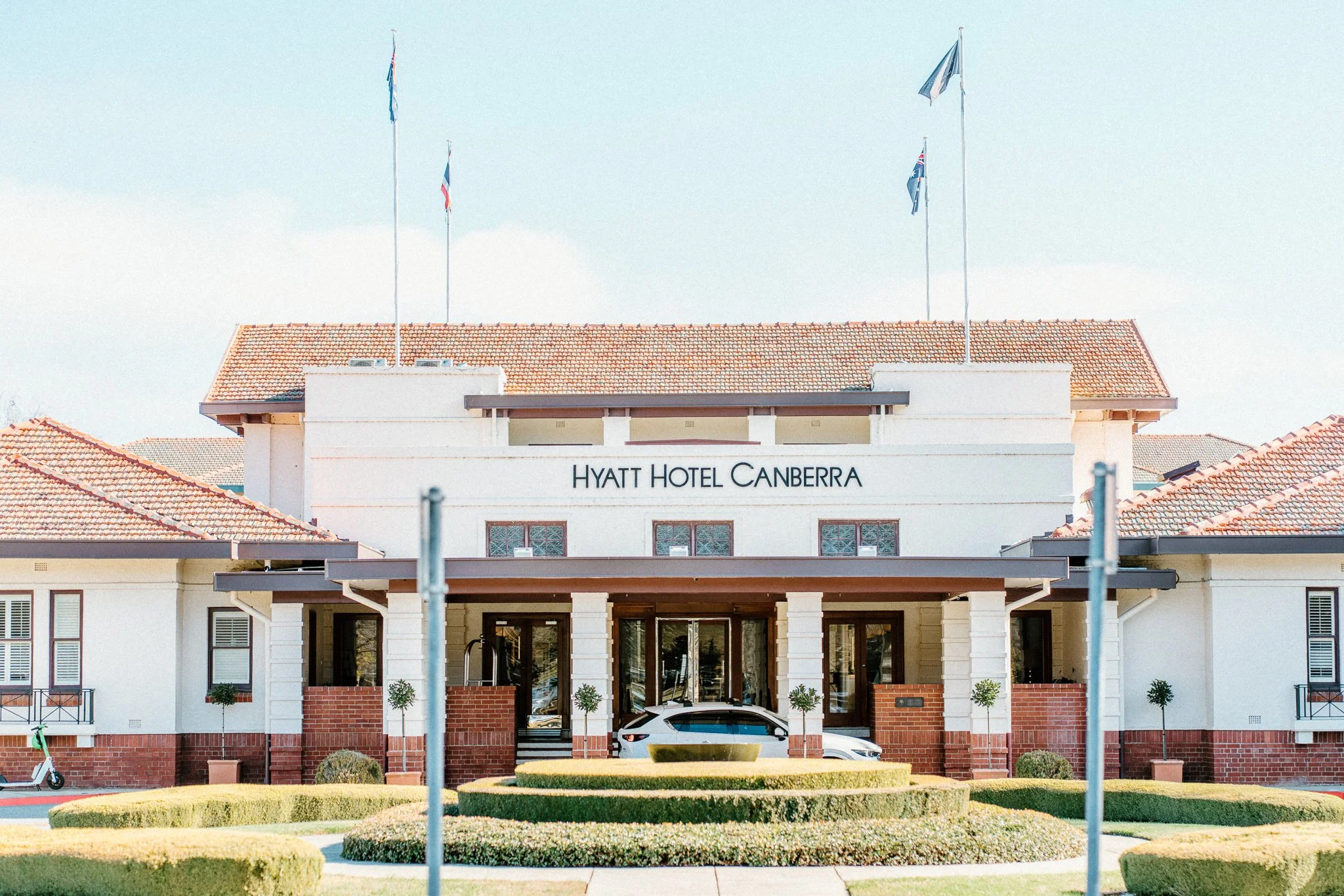 Front view of the Hyatt Hotel Canberra building with a sign, several flags, manicured bushes, a parked car, and a bicycle in the foreground.
