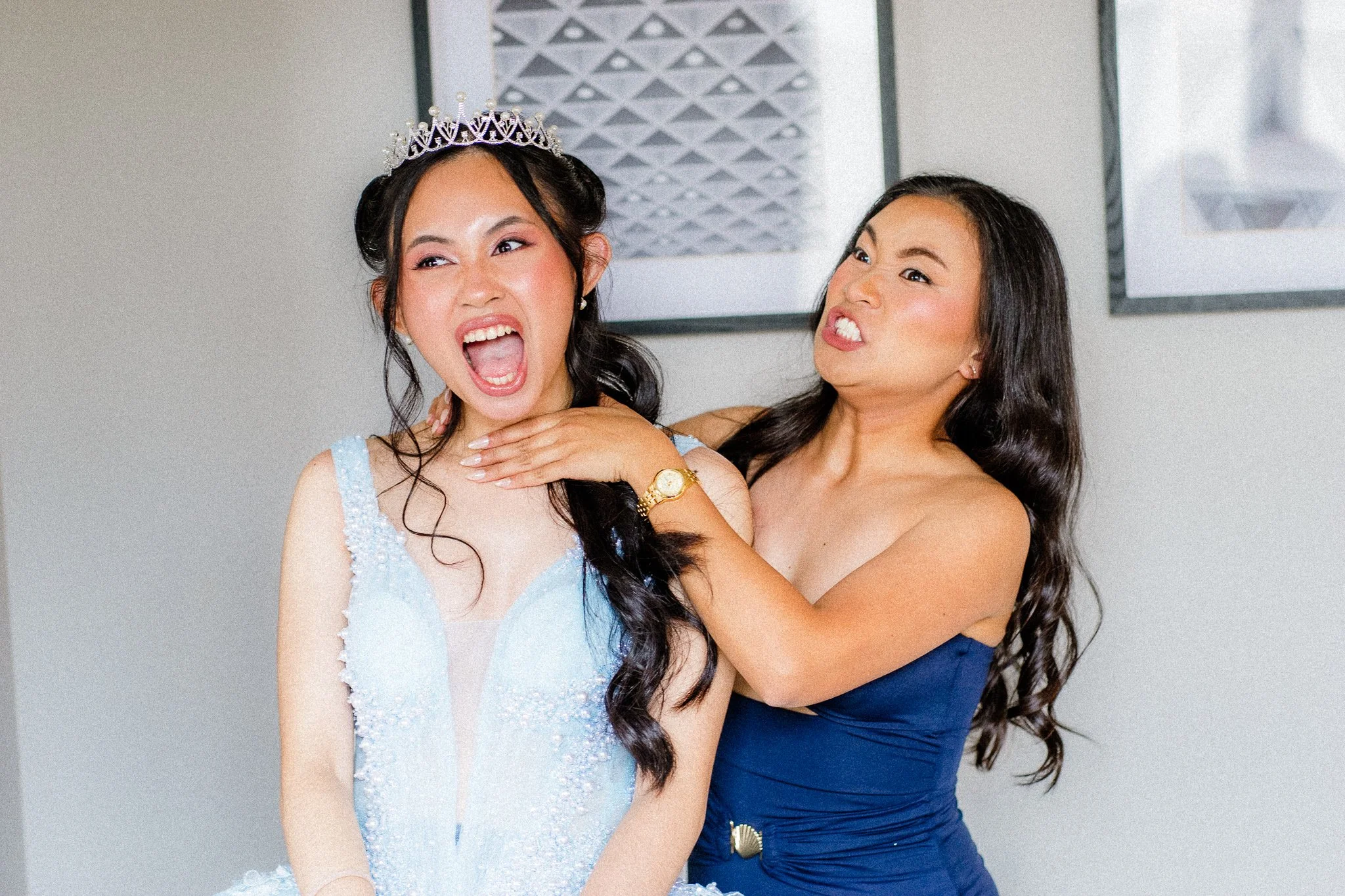 Two women celebrating, one wearing a crown and a light blue dress, the other in a dark blue dress, posing playfully together indoors.