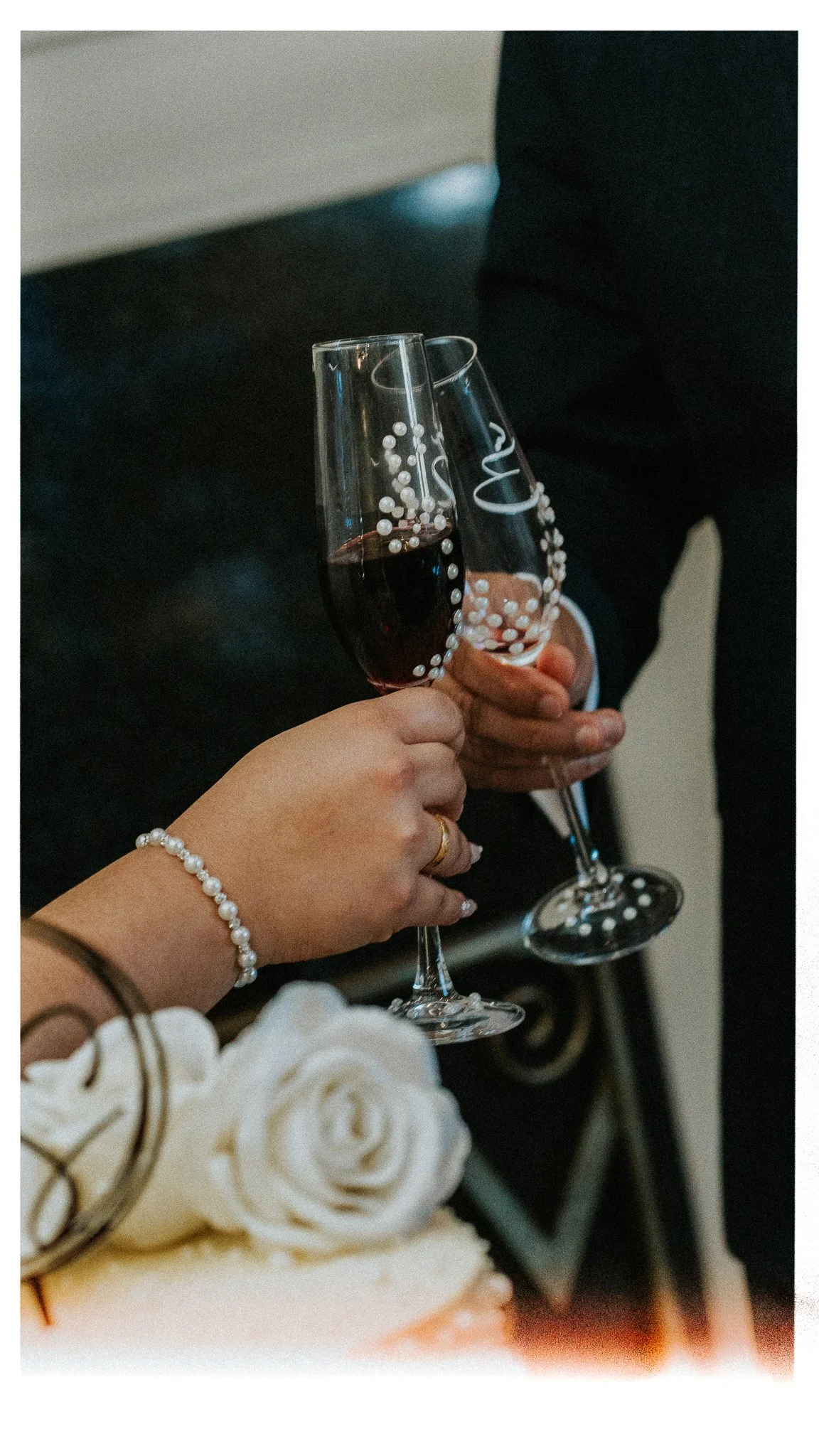 Two people holding glasses of red wine, one is wearing a pearl bracelet and rings, and a bouquet of white roses is visible in the foreground.