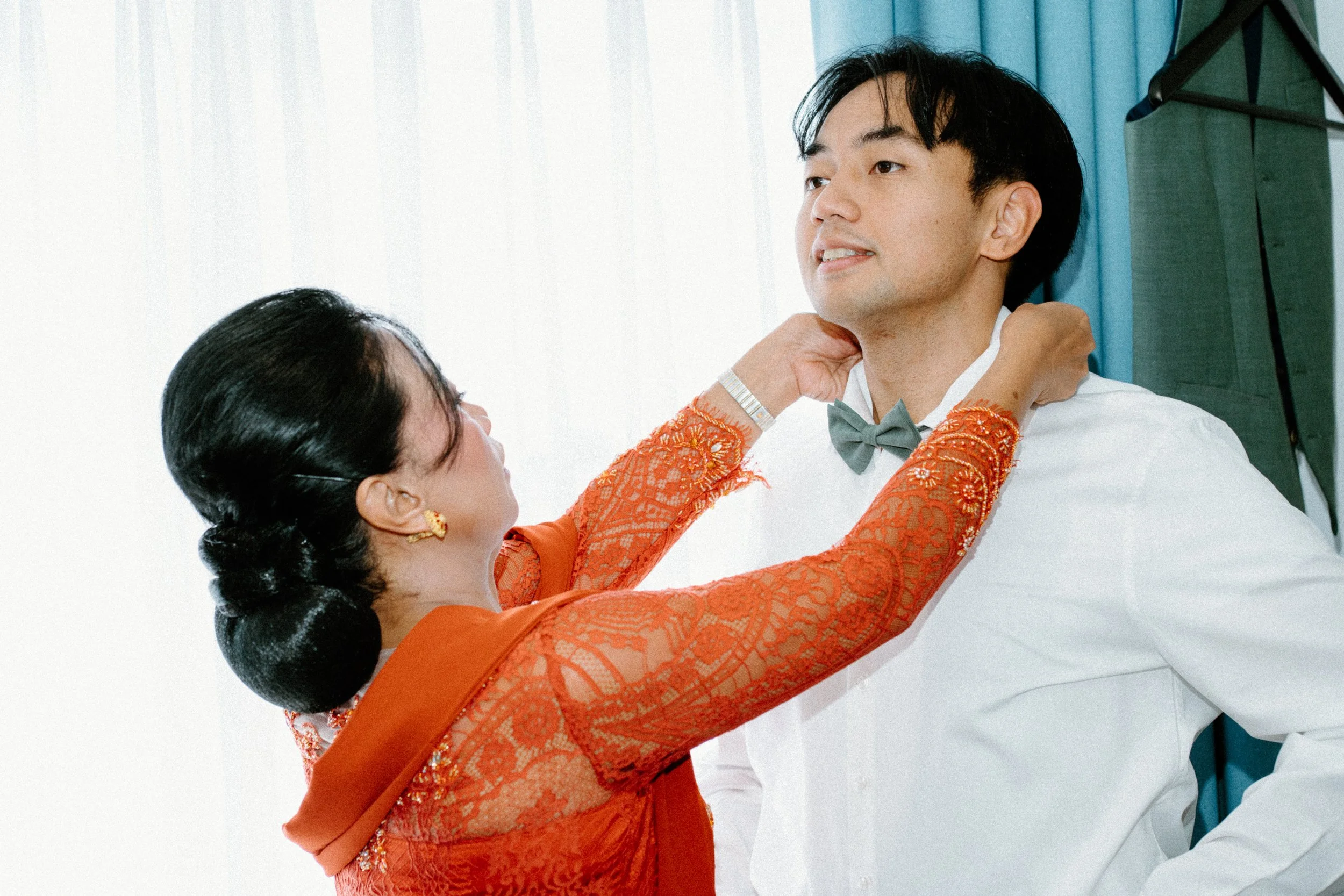 An Indian man dressed in formal attire with a bow tie, appears to celebrate a wedding or cultural event with a woman adjusting his collar, in a bright indoor setting.