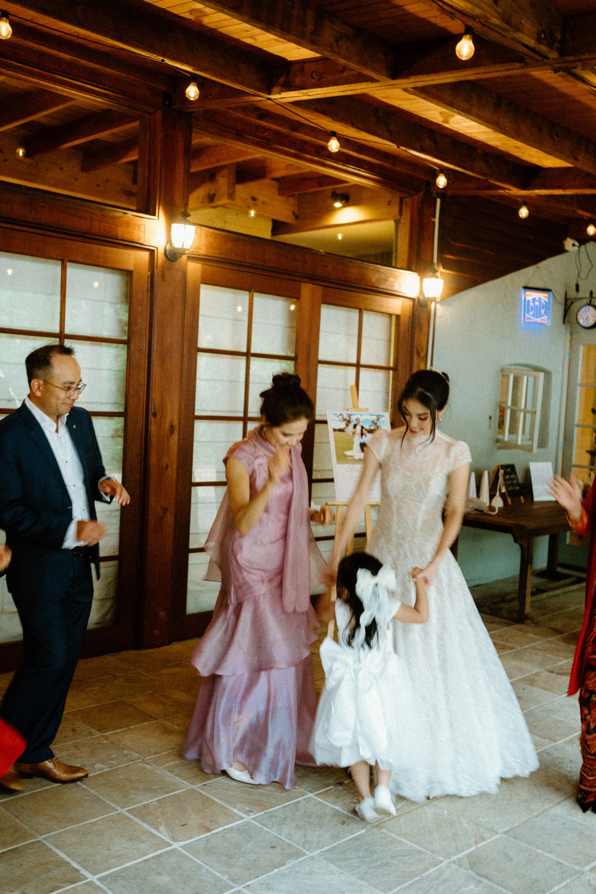 A wedding reception scene with a bride in a white wedding dress dancing with a young girl in a white dress, surrounded by adults, in a warmly lit indoor setting with wooden accents and traditional decor.